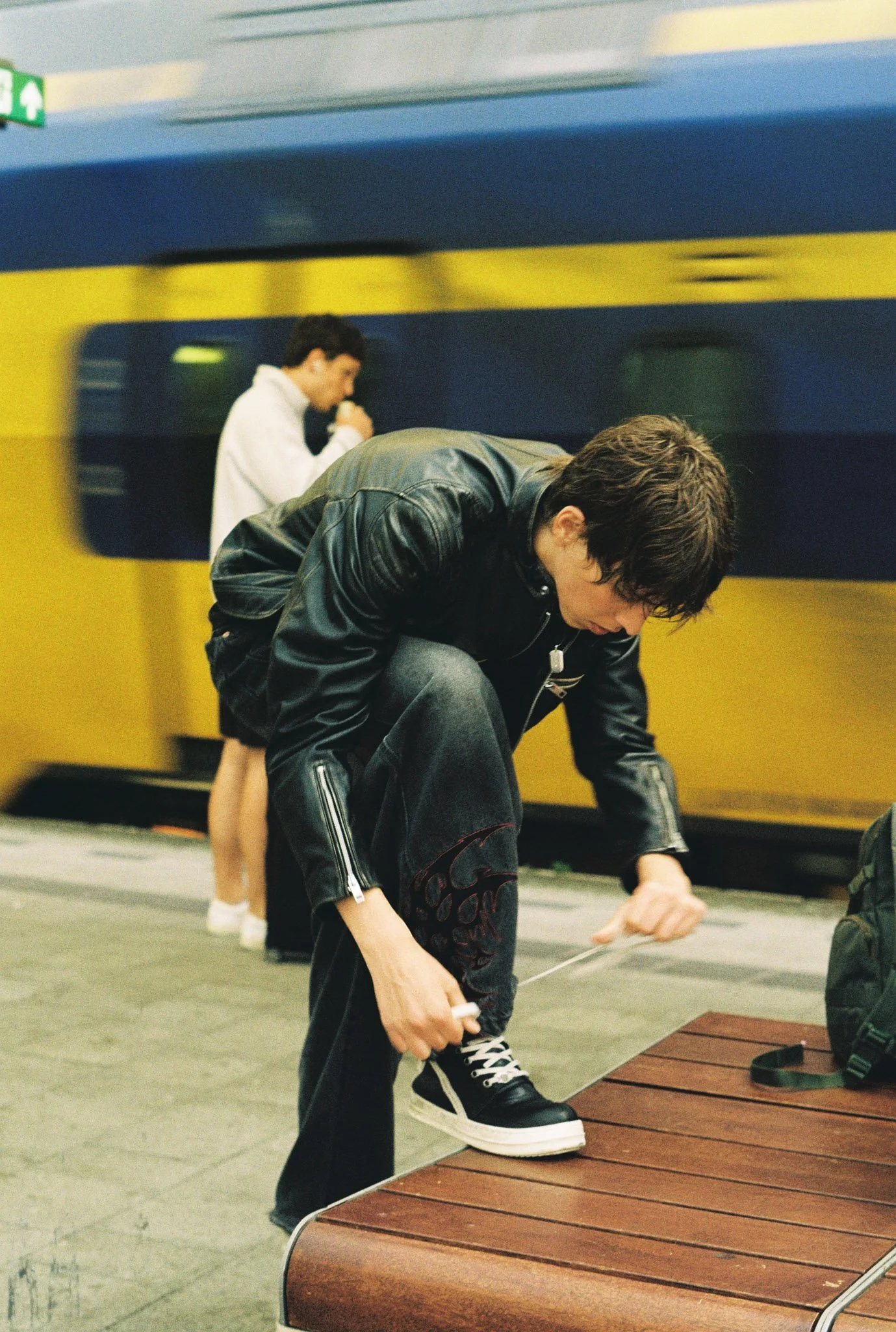 Two young men in black leather jackets are tying their shoes on a wooden bench in a train station. In the background, a young woman in a white jacket and shorts is standing and looking down.