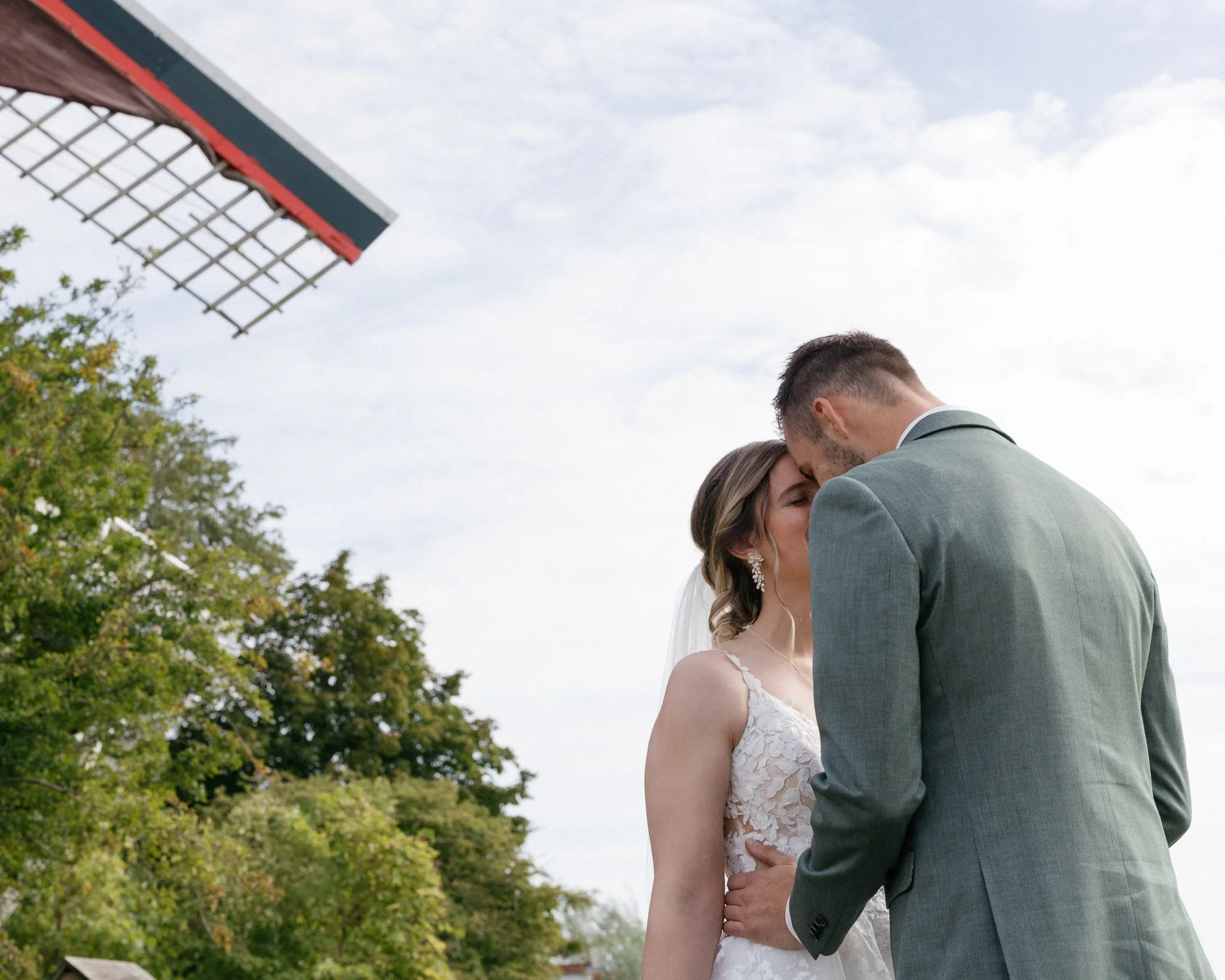 A bride and groom are embracing outdoors, with the groom kissing the bride on the forehead. The bride is wearing a white lace wedding dress and earrings, and the groom is in a gray suit. There are trees and a cloudy sky in the background, and part of a seating area or structure is visible at the top left corner.