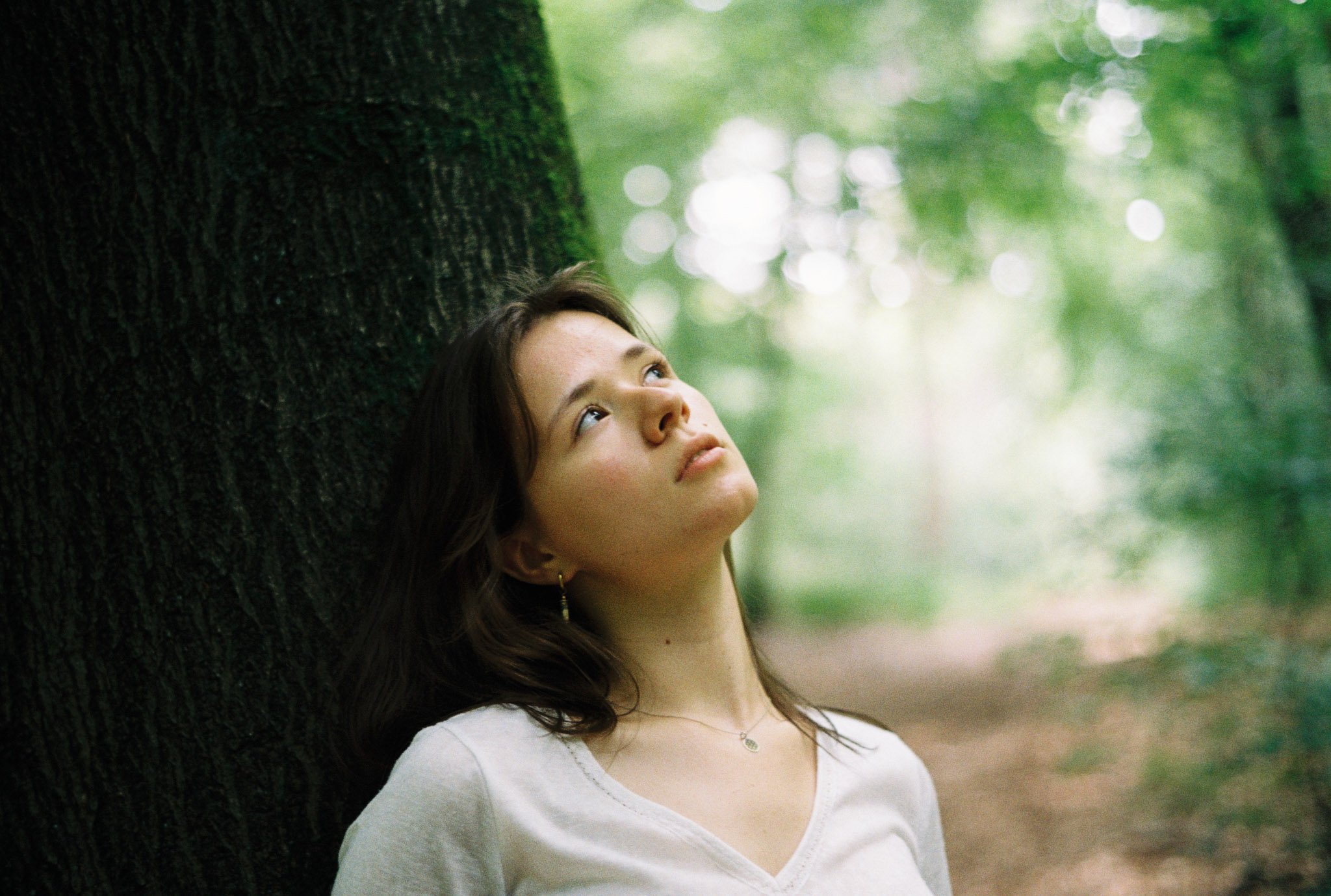 A young woman with dark hair and light skin looks upwards while leaning against a large tree in a green forest, with blurred foliage in the background.