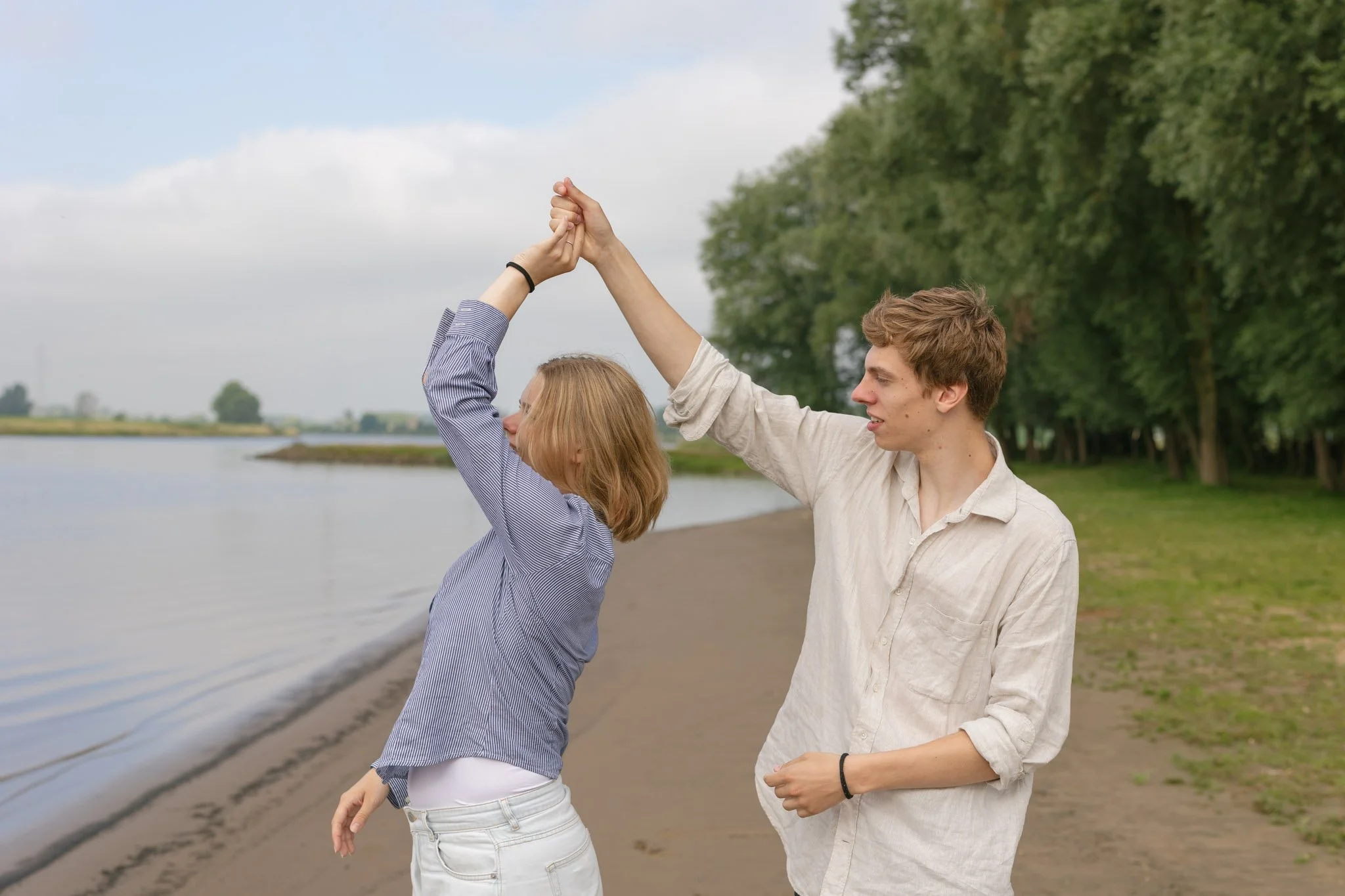 A young man and woman high-five each other by a body of water in a park, with trees and a cloudy sky in the background.