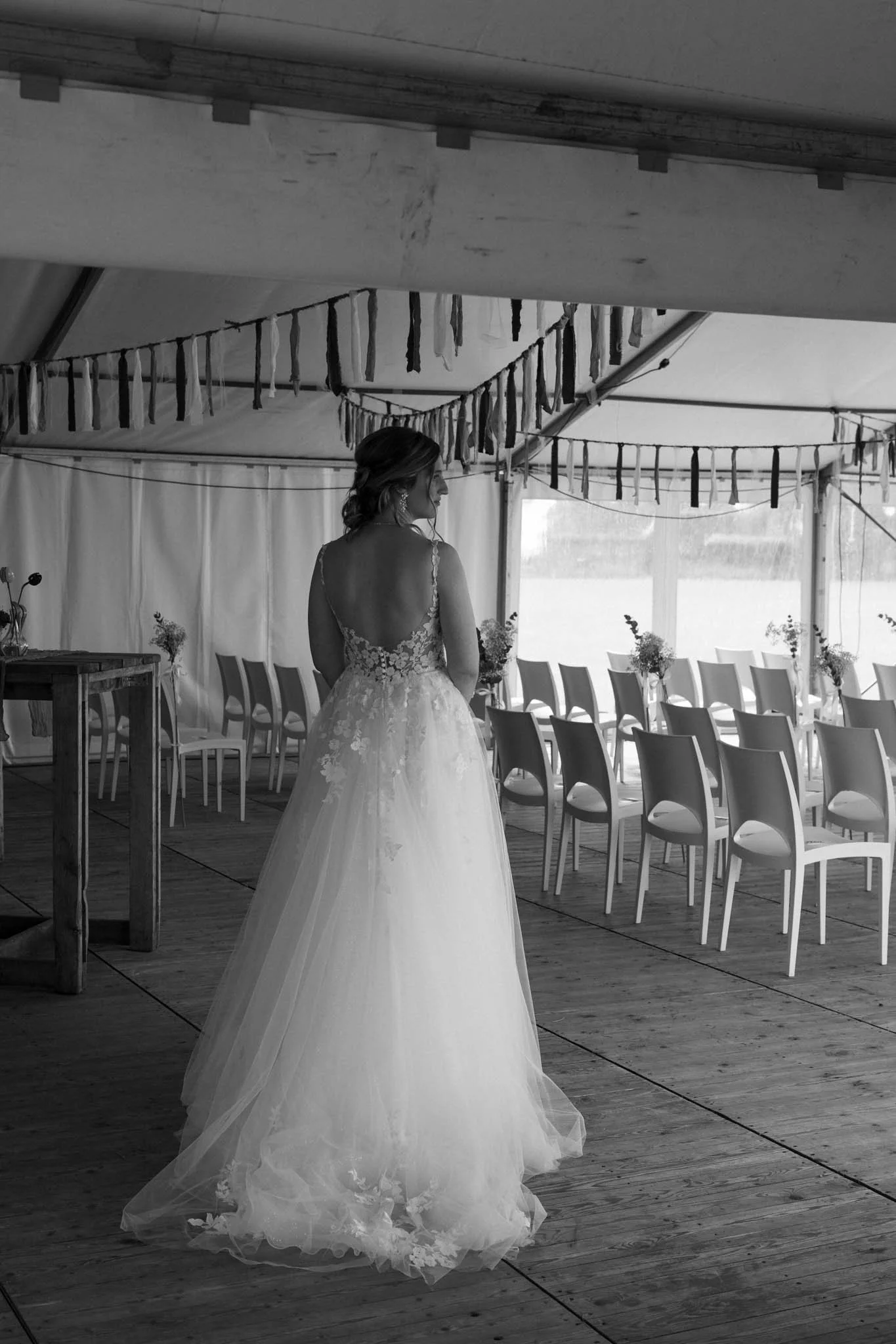 A woman in a wedding dress standing alone in an indoor event space, with rows of chairs and decorative hanging fabric strips overhead.