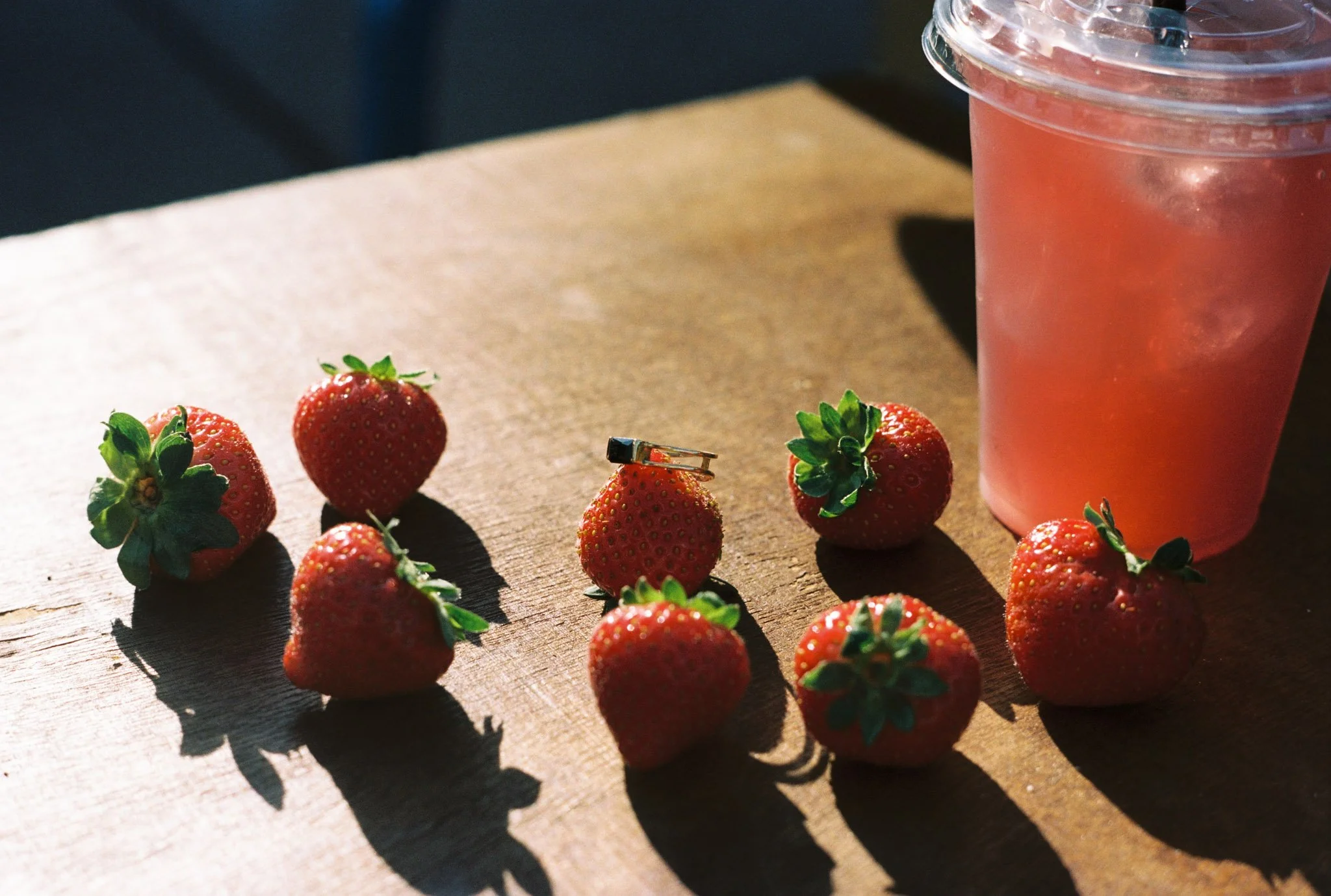 Fresh strawberries scattered on a wooden surface with a pink drink in a plastic cup on the right side.