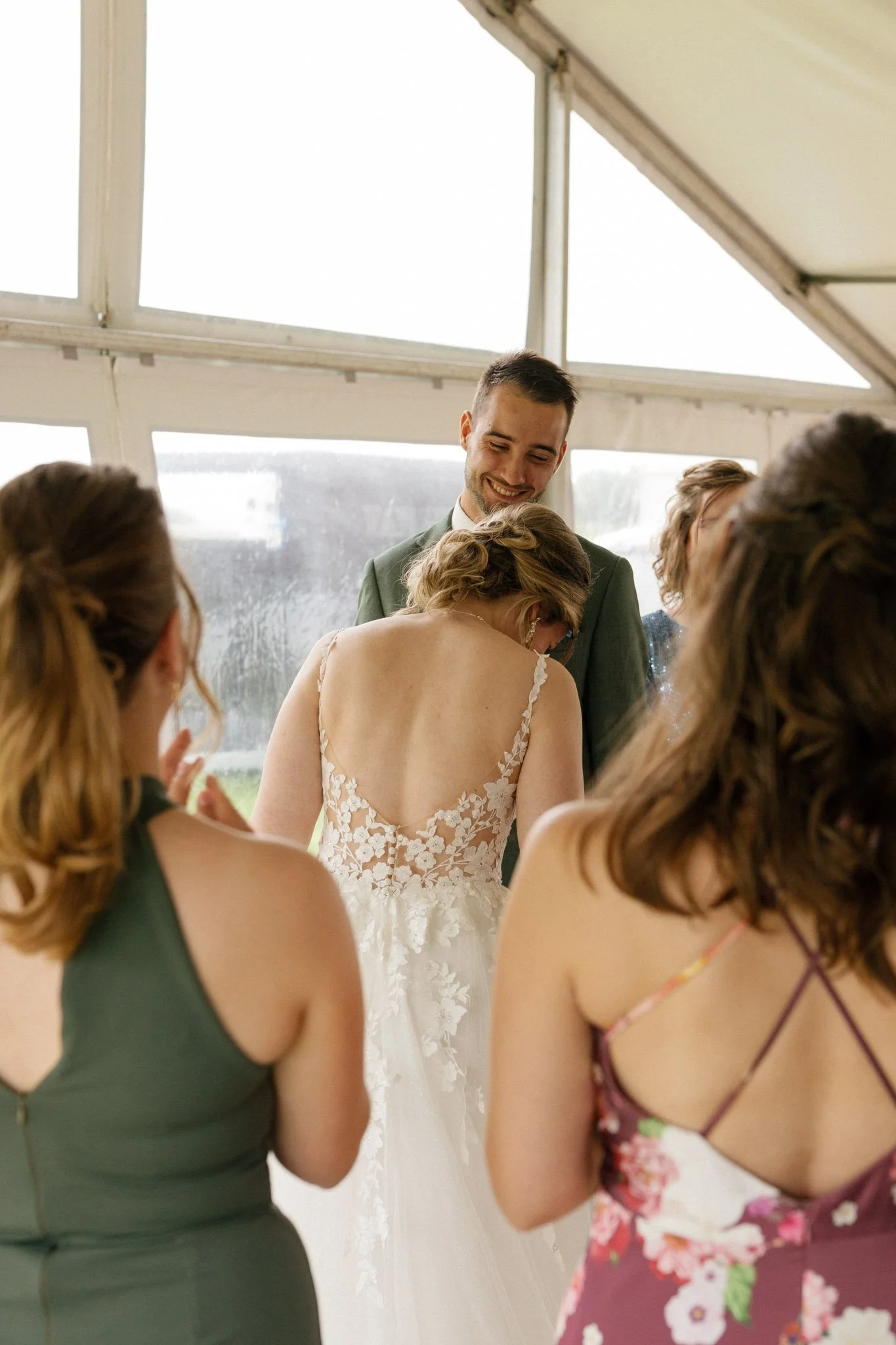 People at a wedding celebration, with one couple embracing and smiling, and others around them in a glass-walled venue.