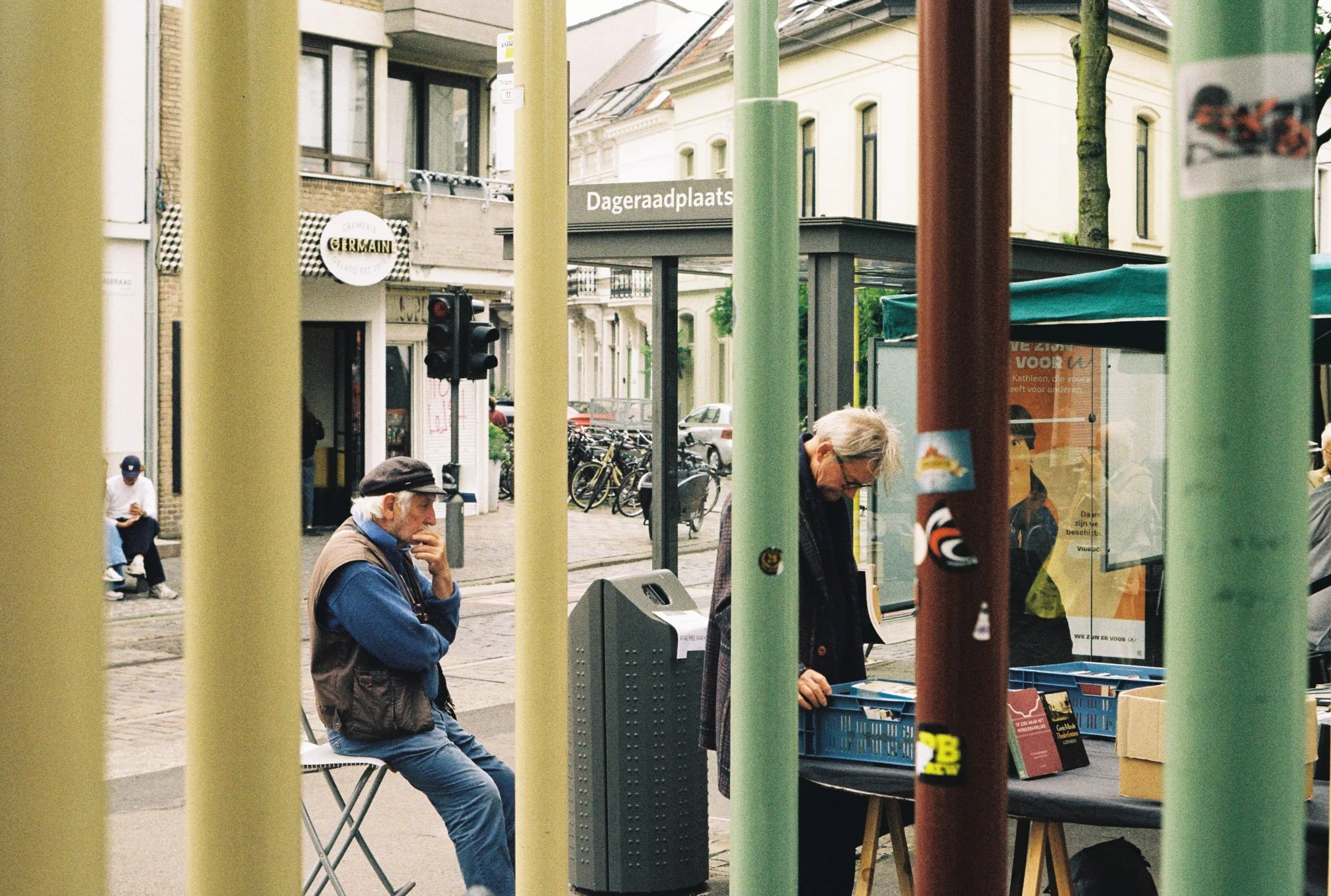 People waiting at a street-side kiosk with colorful vertical poles in the foreground, some sitting and some standing, in an urban area with buildings and bike racks visible in the background.
