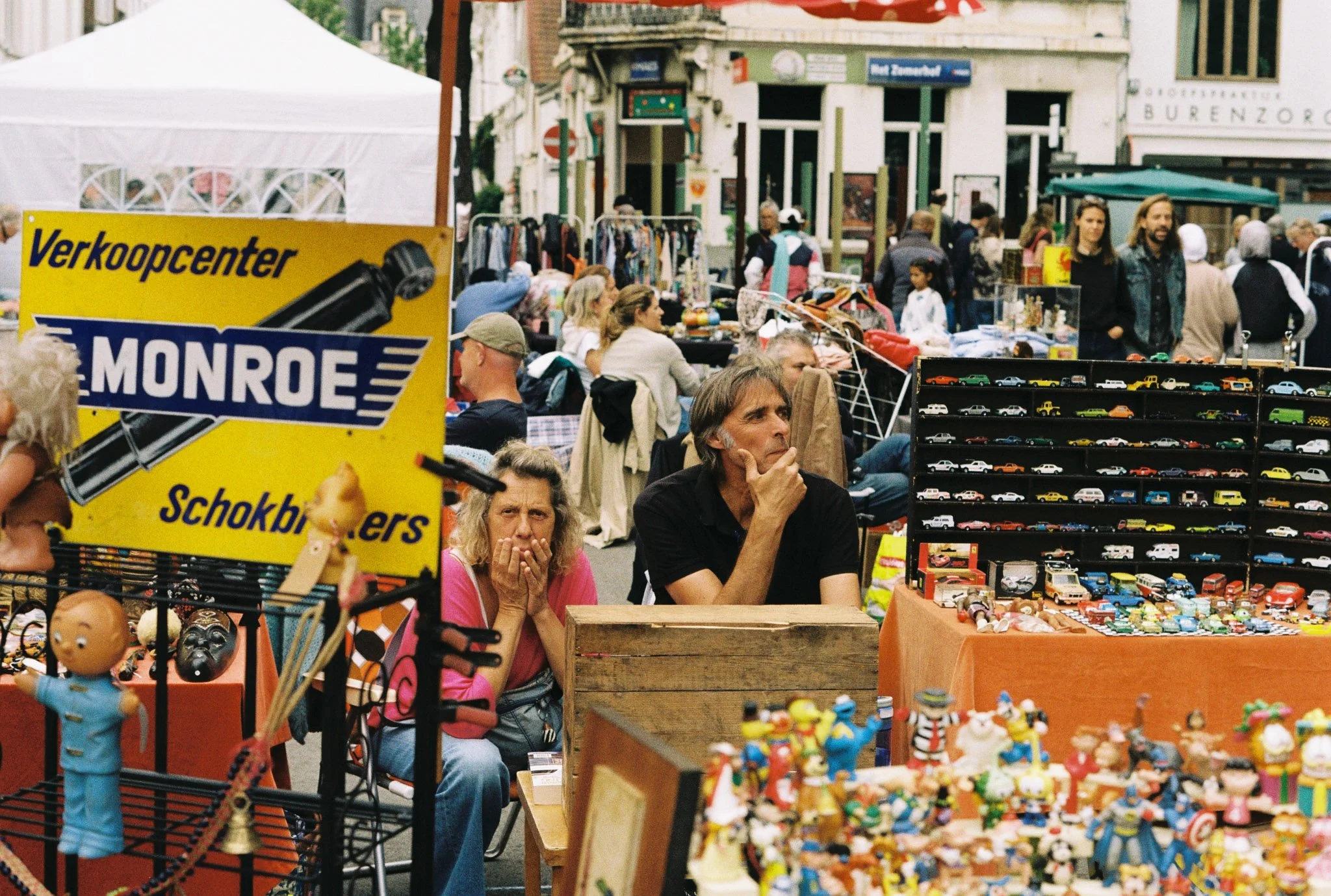 A busy street market with vendors selling toys, clothing, and collectibles, surrounded by shoppers and pedestrians.