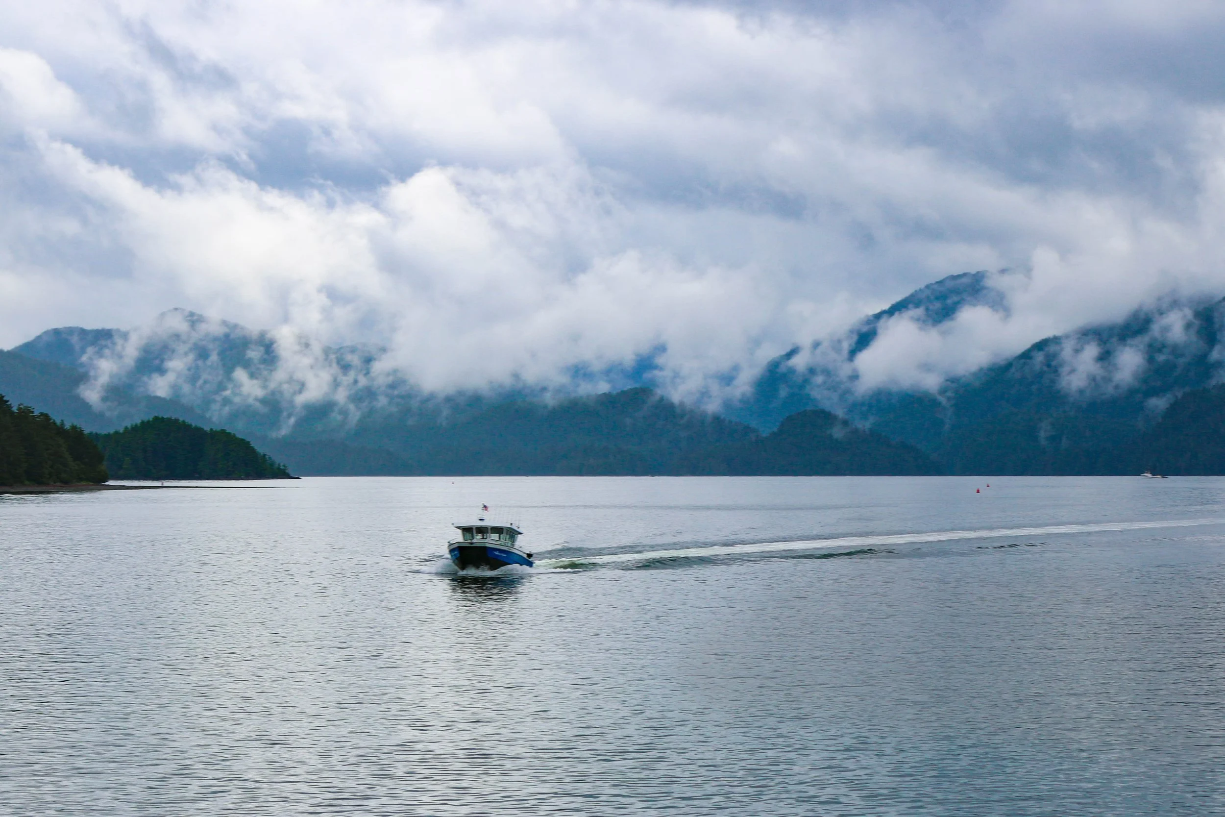 A boat traveling on a calm body of water, with lush green hills and fog-covered mountains in the background, and cloudy sky above.