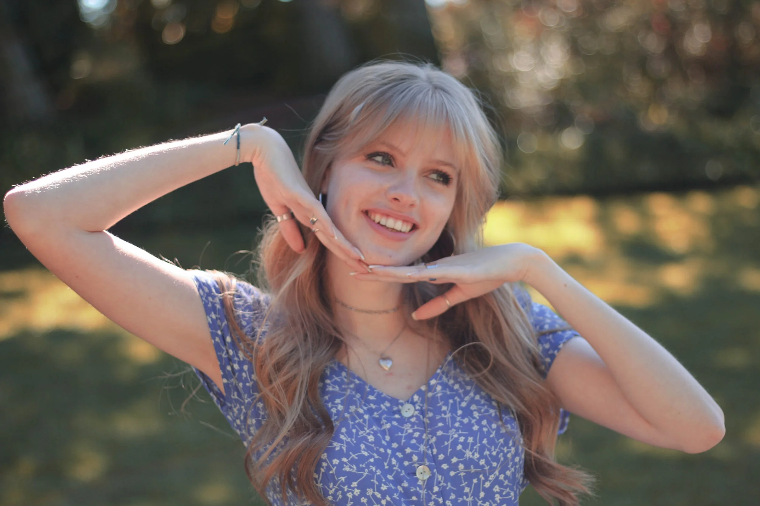 A young woman with long, wavy, blonde hair wearing a blue dress with white floral patterns, smiling and posing outdoors with her hands under her chin against a background of blurred trees and sunlight.