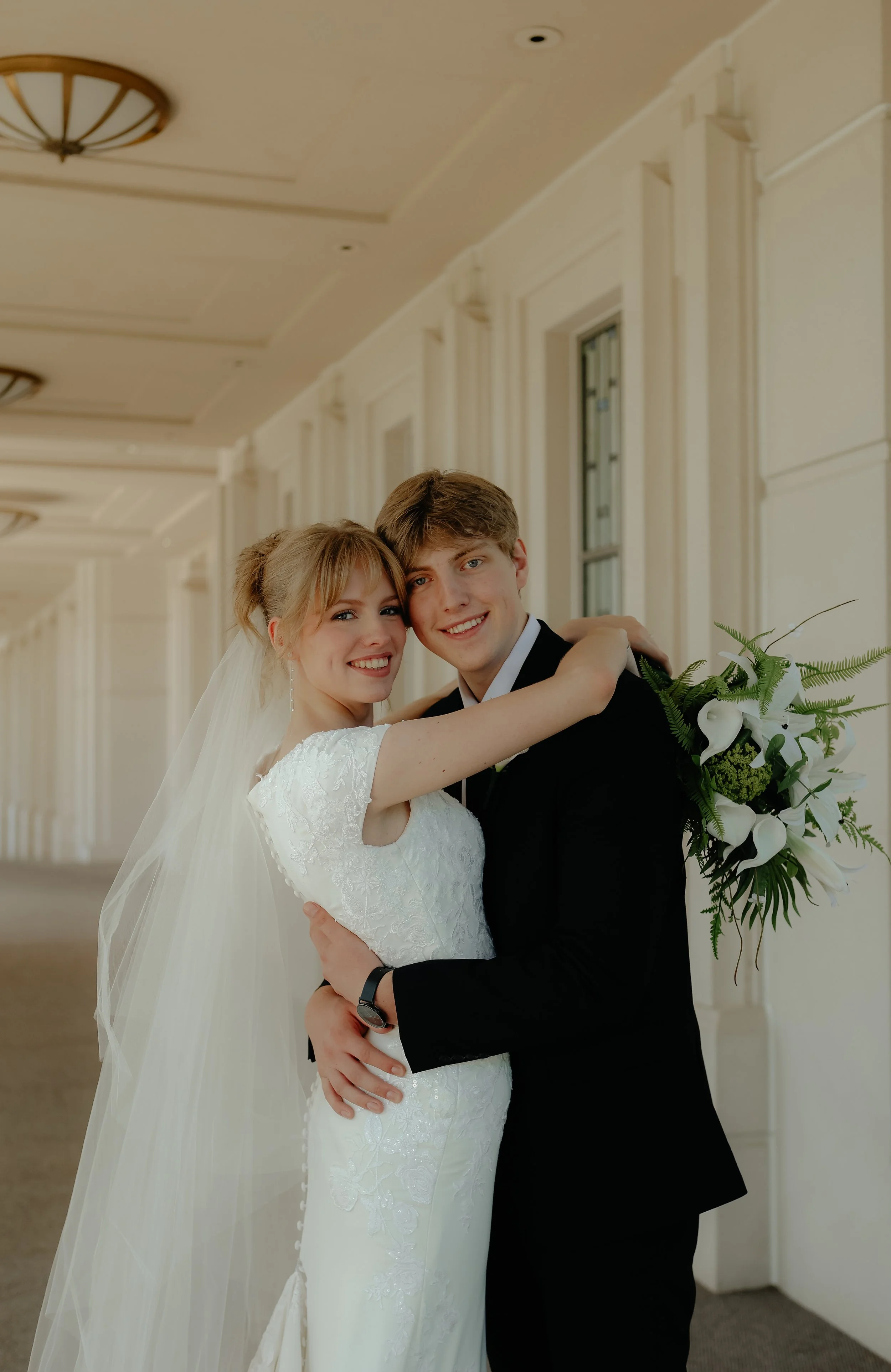 A bride and groom smiling and hugging at their wedding ceremony, with the bride holding a bouquet of white calla lilies and greenery in a bright, elegant corridor.