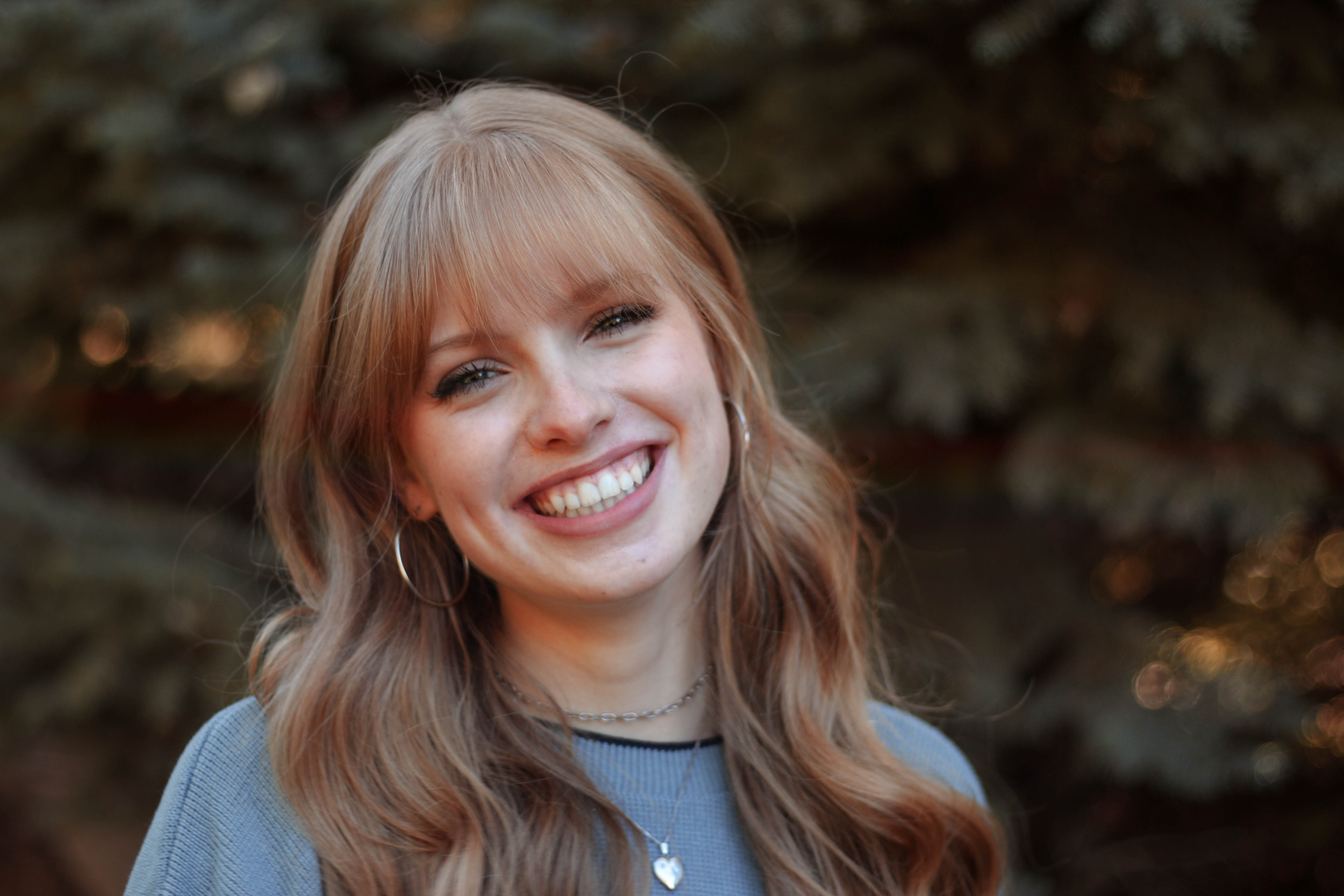 Smiling young woman with blonde hair, wearing hoop earrings, a necklace with a heart pendant, and a blue sweater outdoors with blurred trees in the background.