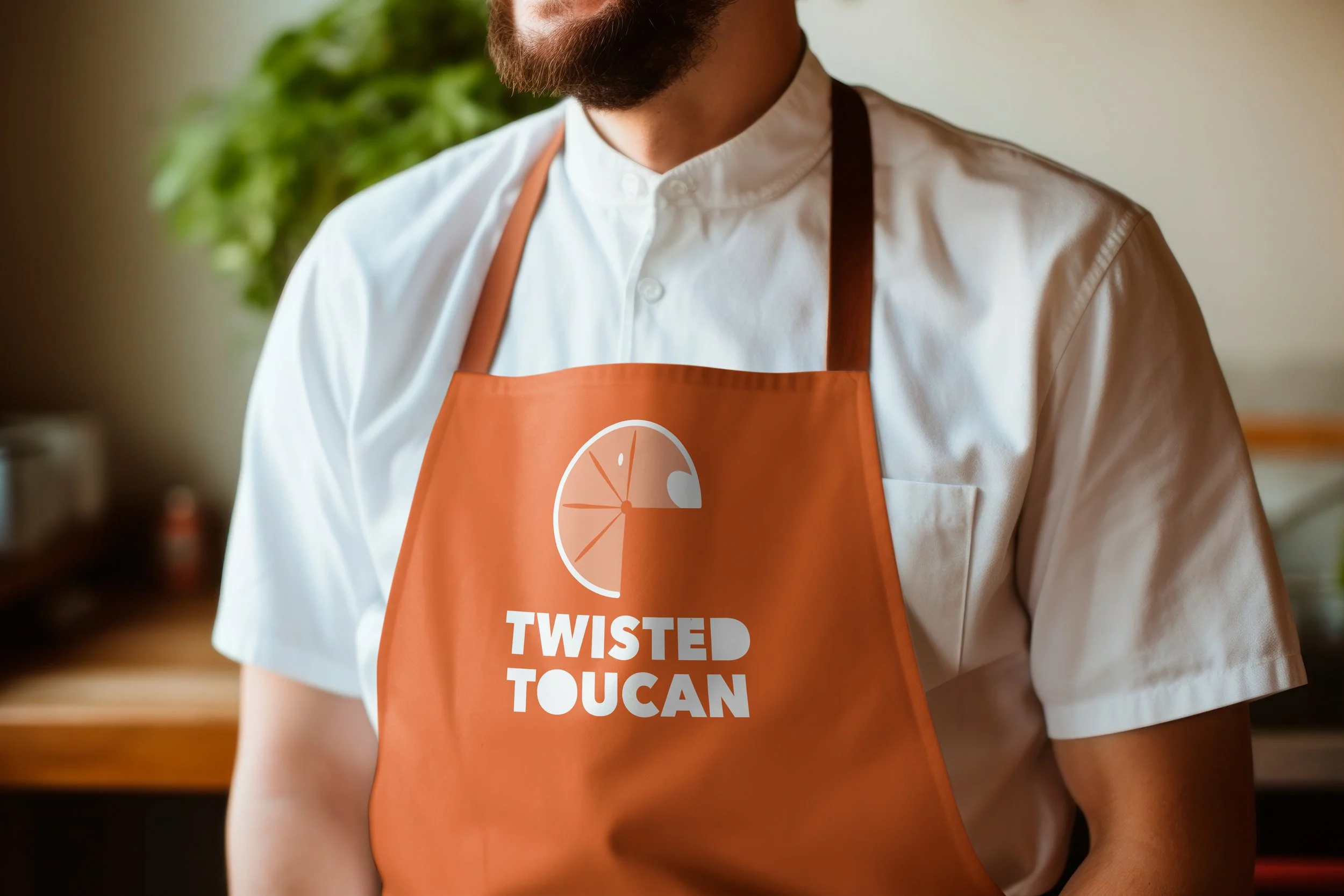 Close-up of a man wearing a white chef's coat and an orange apron with the words "Twisted Toucan" and a logo of a sliced orange. The man has a full beard and is smiling, with a green plant in the background.