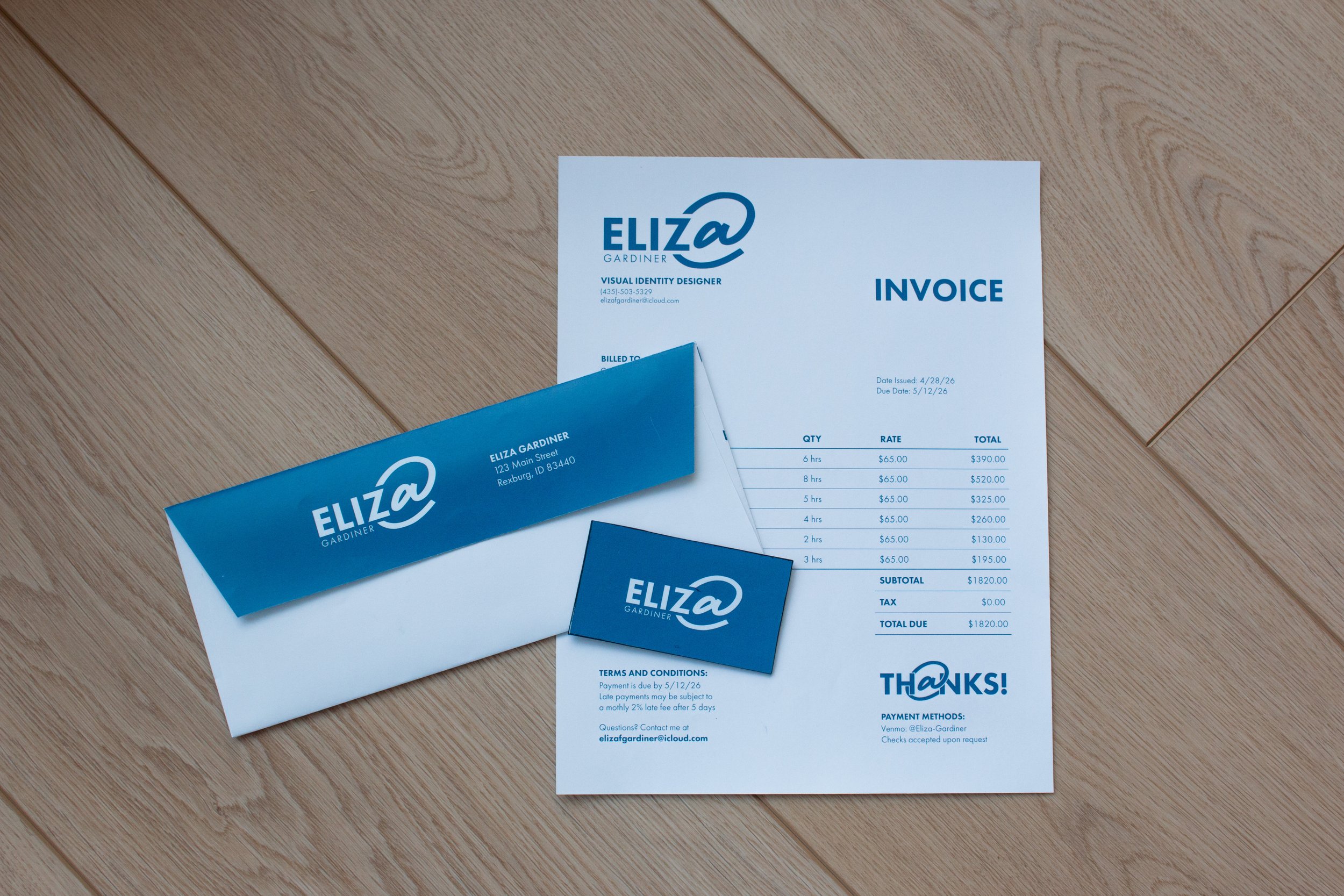 An invoice, thank you card, a business card, and two blue paper clips all featuring the name and logo of Eliza Gardiner, a visual identity designer, on a wooden surface.