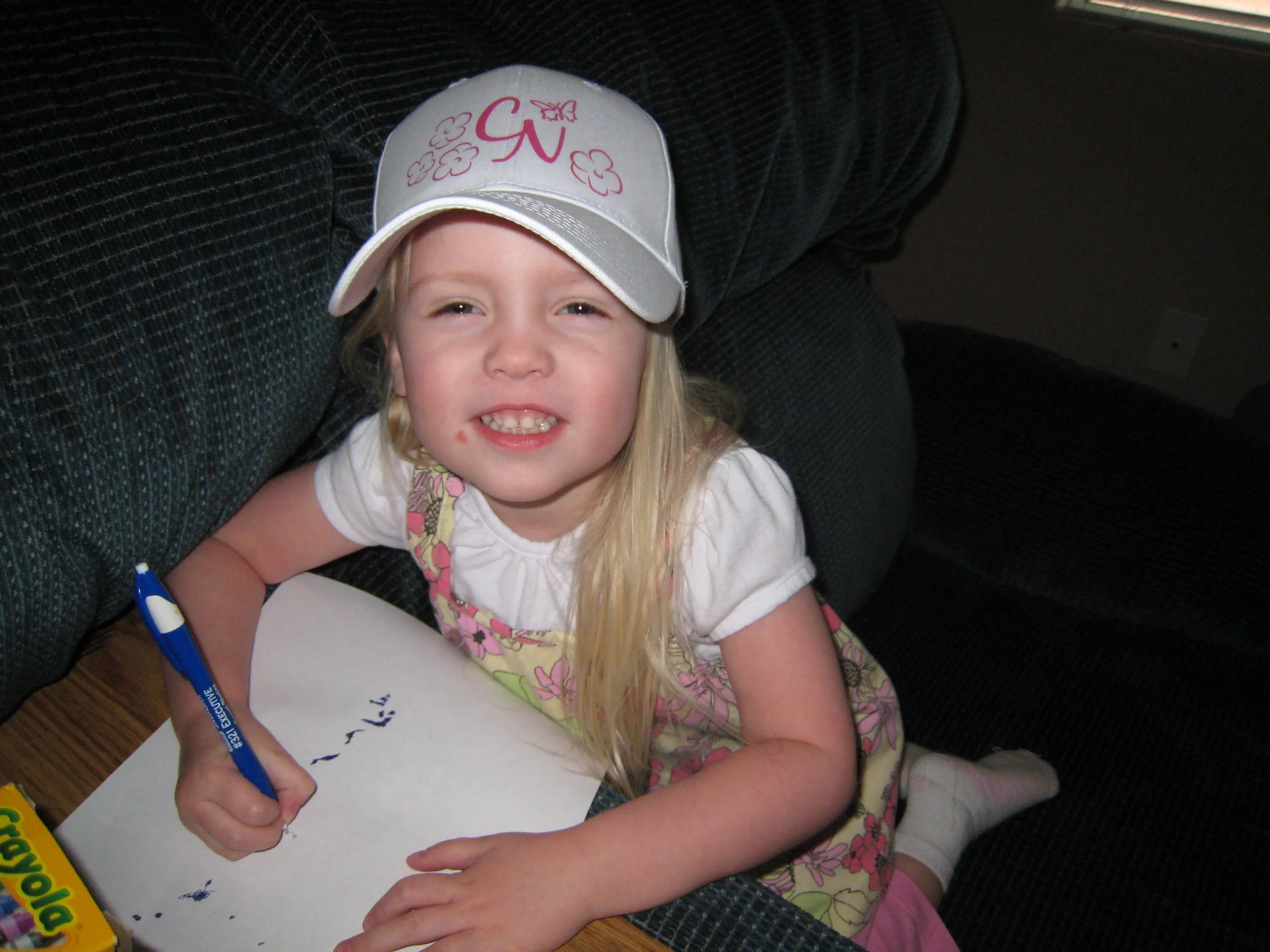 A young girl with blonde hair wearing a white hat with pink flowers and a butterfly, smiling and sitting at a table, writing on a piece of paper with a blue marker.