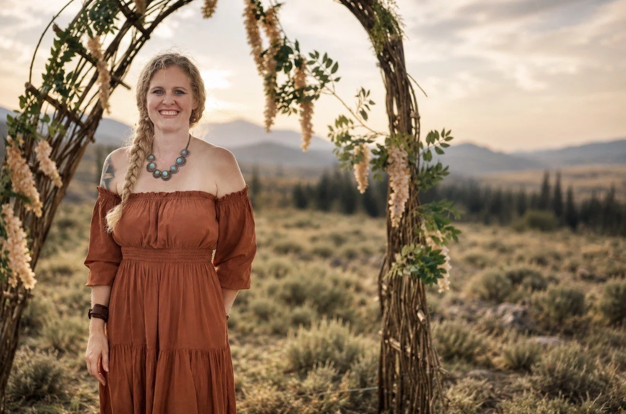 A woman with blonde hair in a braid, wearing an off-the-shoulder rust-colored dress and a necklace, standing outdoors on a grassy plain at sunset, framed by a wooden arch decorated with flowers and green foliage.