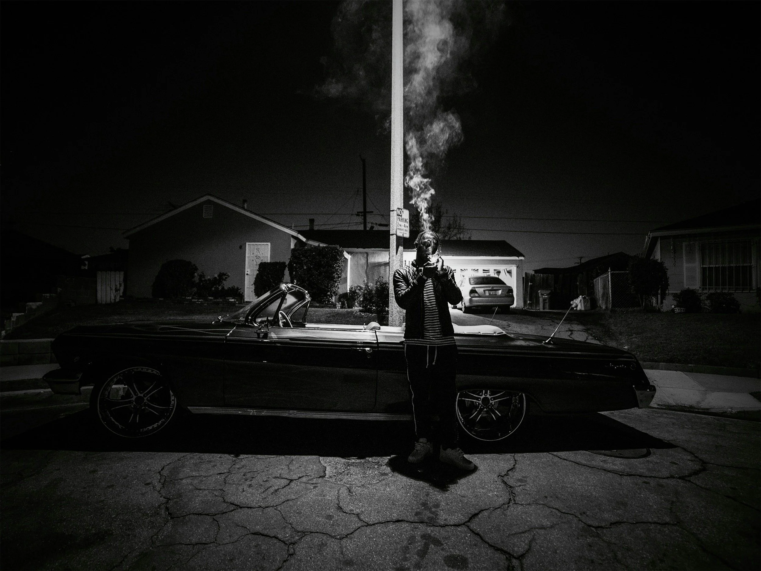 A person standing in front of a black vintage convertible car at night, smoking a cigarette with smoke rising up, on a cracked asphalt driveway with houses and a streetlight in the background.