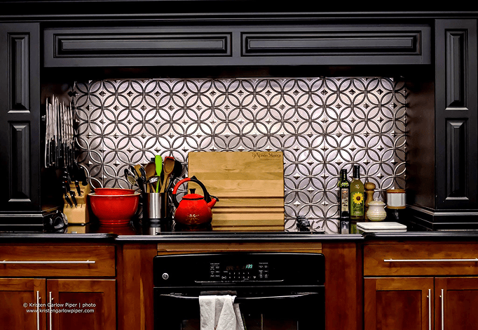 Apron Strings Store kitchen counter with knife blocks, bowls, utensils, cutting board, cooking oils, and small containers, set against a patterned backsplash and black cabinetry.