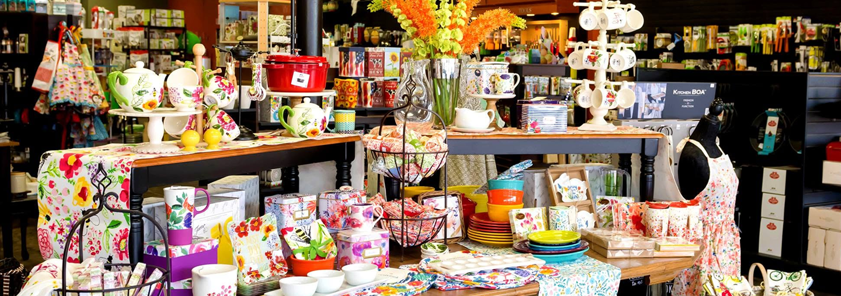 Display of colorful kitchenware and decorative items at Apron Strings Kitchen store, including mugs, bowls, plates, floral-patterned teapots, and a mannequin with a floral apron, arranged on tables and shelves.