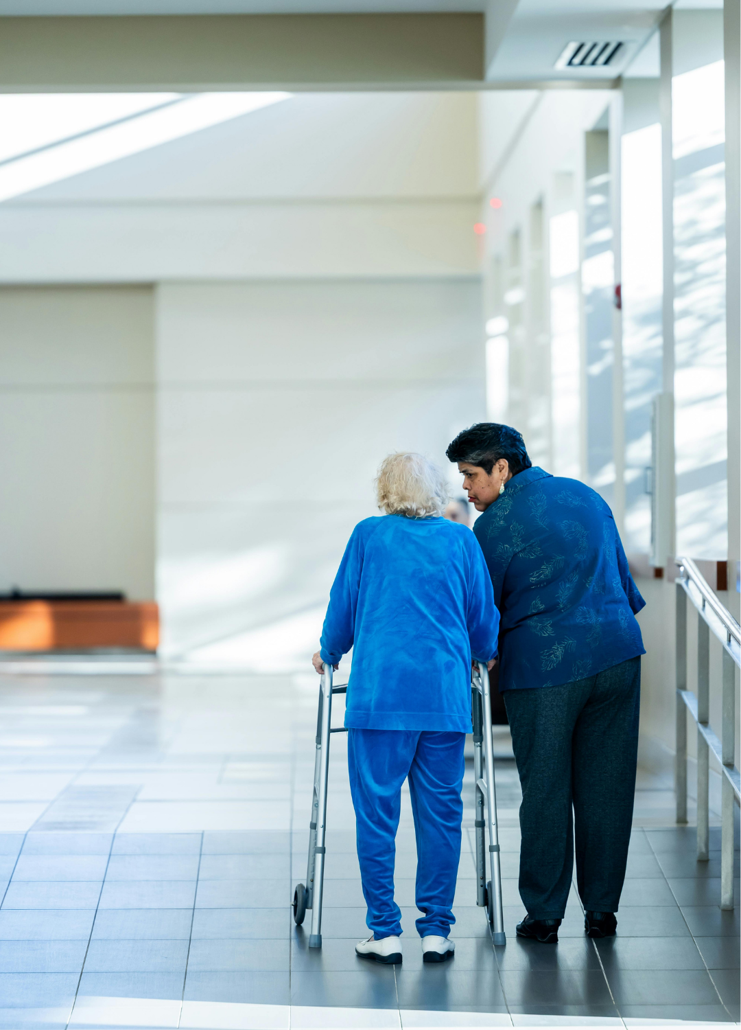 An elderly woman in a blue outfit using a walker and a caregiver walk together down a hospital corridor, engaged in conversation.