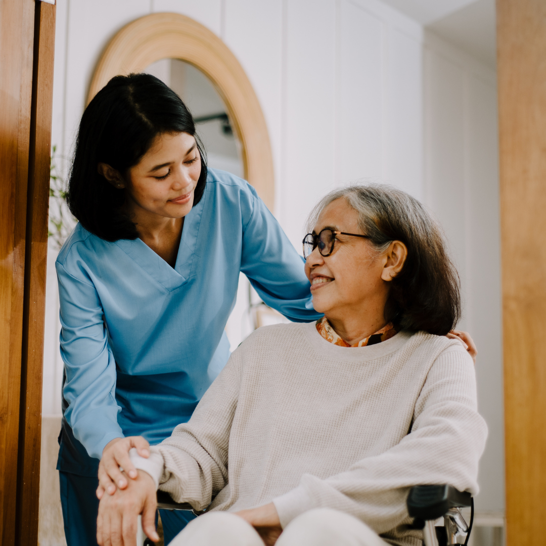 Nurse in blue scrubs smiling and holding the hand of an elderly woman with glasses sitting in a wheelchair.