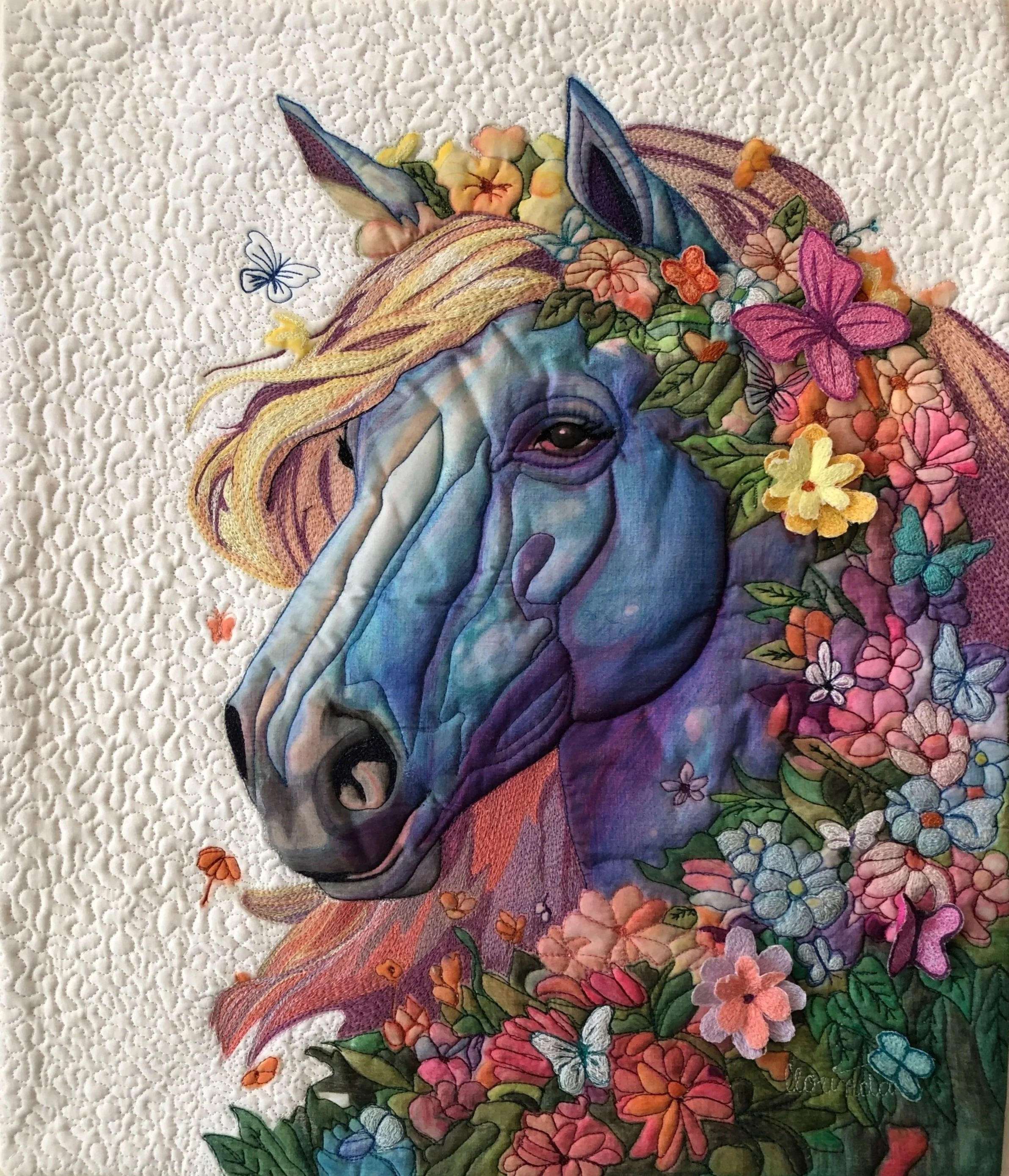 Free-motion Embroidery artwork of a blue horse with a colorful mane, surrounded by flowers and butterflies, on a white quilted background.