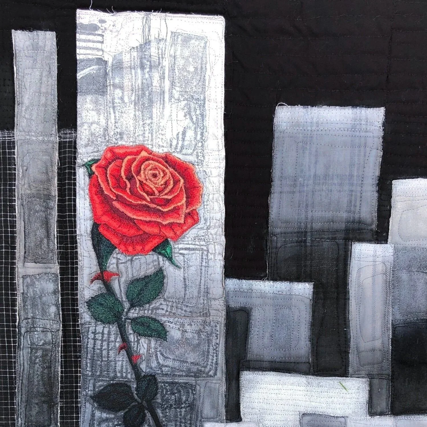 Textile with thread painted red rose and green leaves design, surrounded by gray, black, and white patchwork patterns.