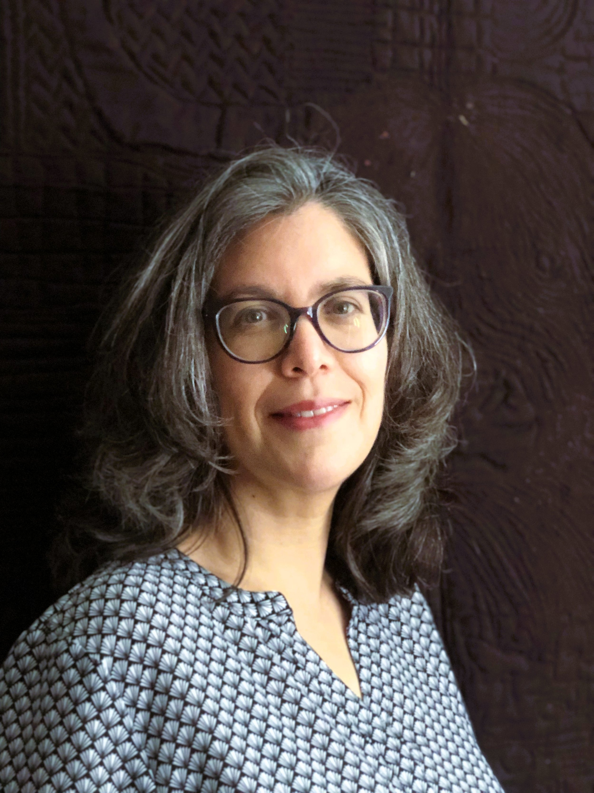 A woman with gray hair and glasses smiling in front of a dark textured background.