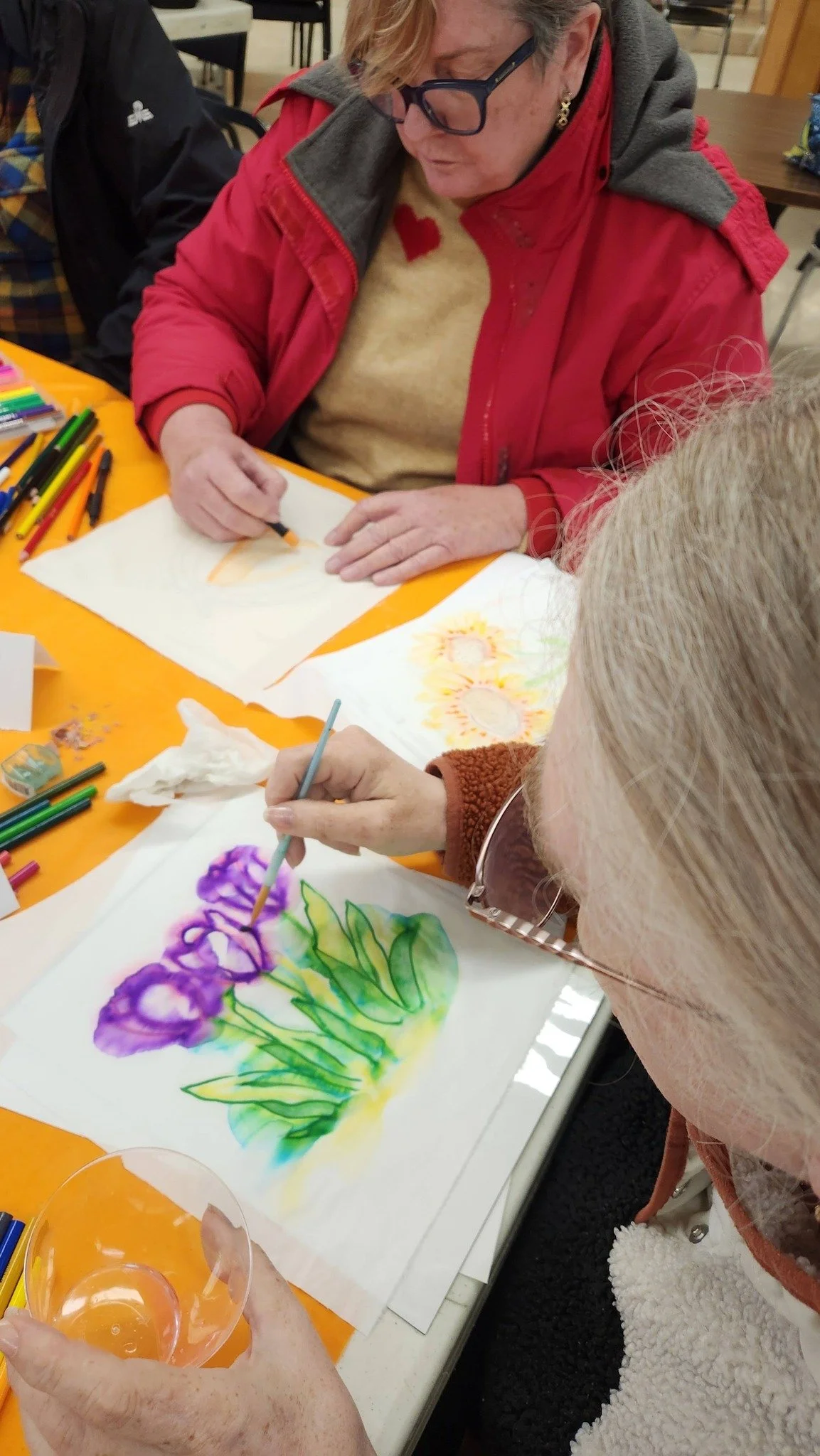 Two women are sitting at a table painting watercolor pictures of flowers. One woman is in a red jacket and the other is in a beige sweater. Art supplies such as markers and paintbrushes are on the table.