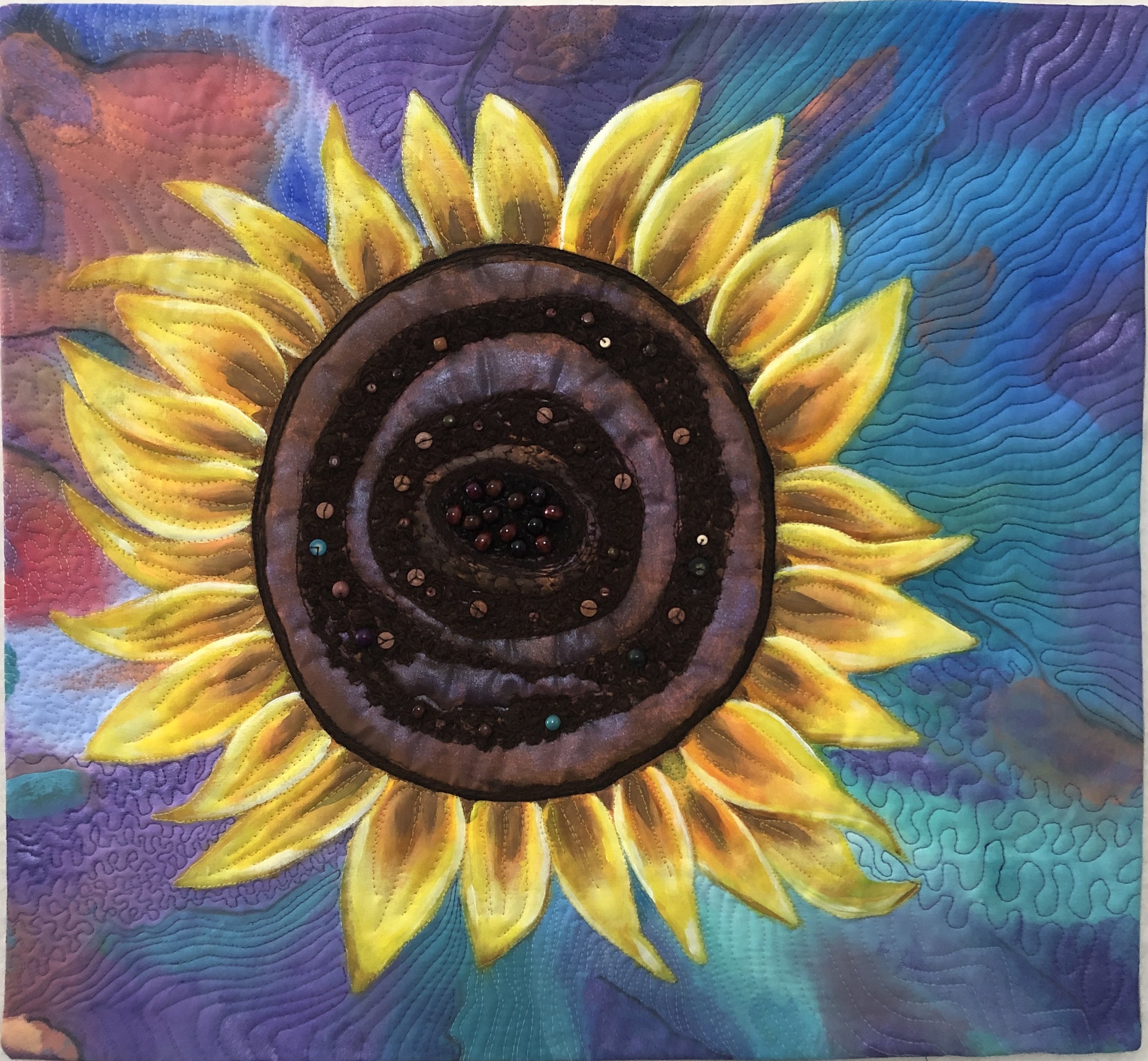 A quilted fabric artwork featuring a sunflower design with bright yellow petals, brown and beige center, and colorful background with purple, blue, and pink hues.