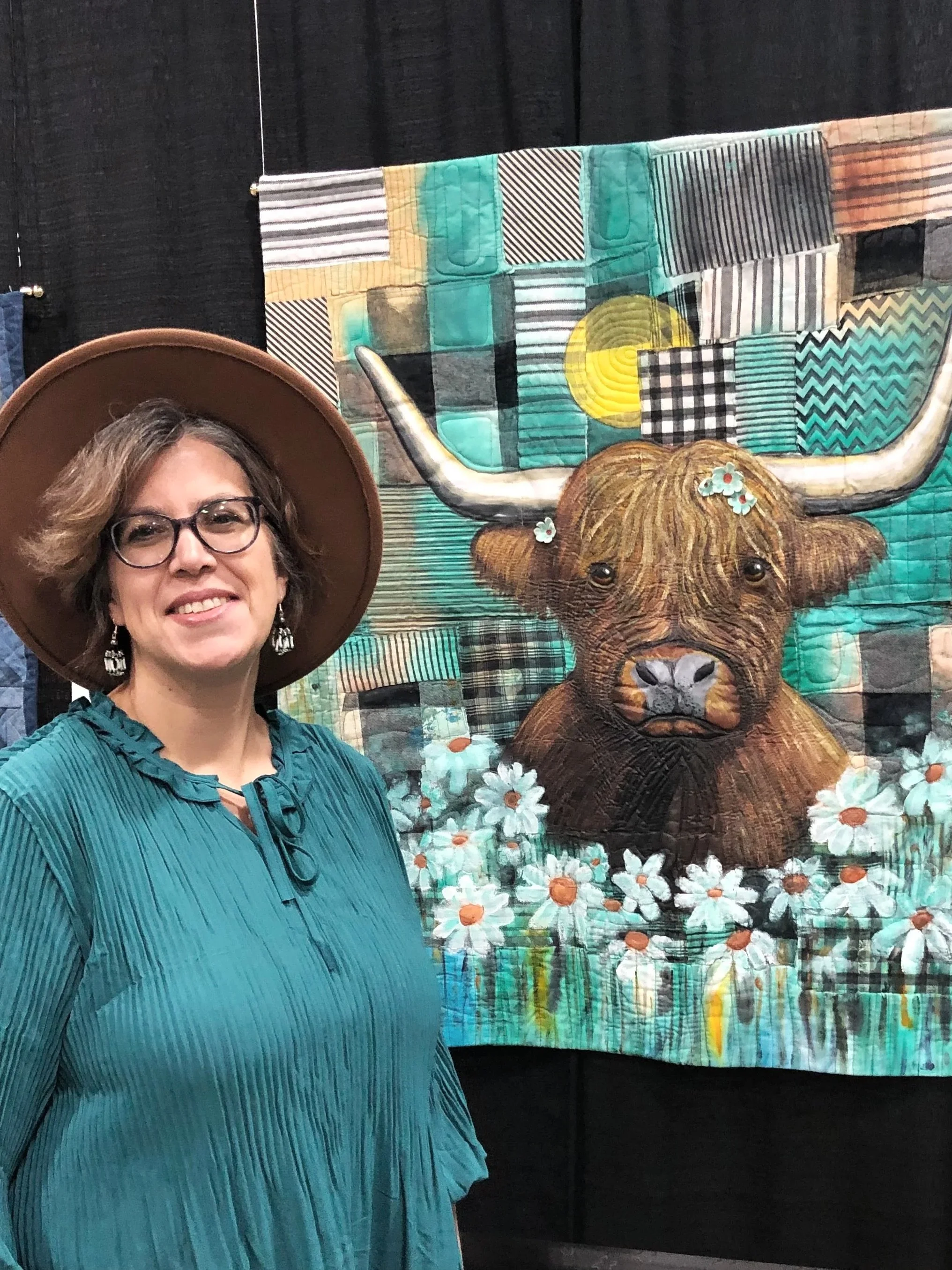 A woman with glasses and a wide-brimmed hat stands in front of a colorful quilted artwork of a bison head with flowers and geometric patterns.