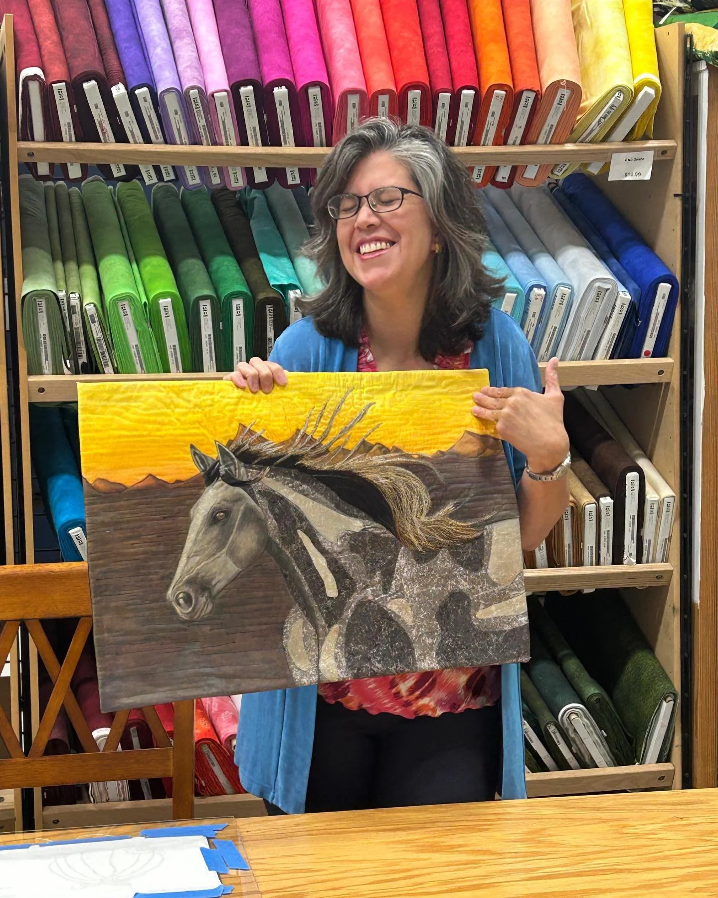 A woman with glasses and gray hair smiling while holding a painting of a horse with a mountainous background. She is standing in front of a shelf filled with colorful rolls of fabric.