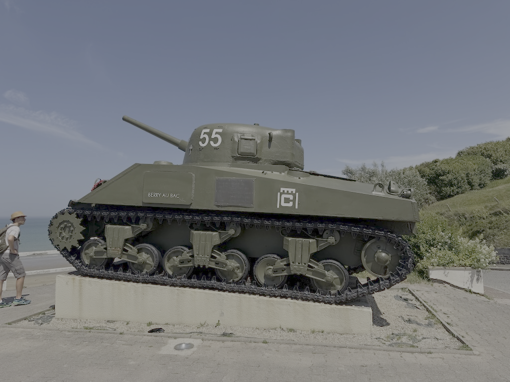 A military tank mounted on a concrete base outdoors with a person walking nearby, under a partly cloudy sky.