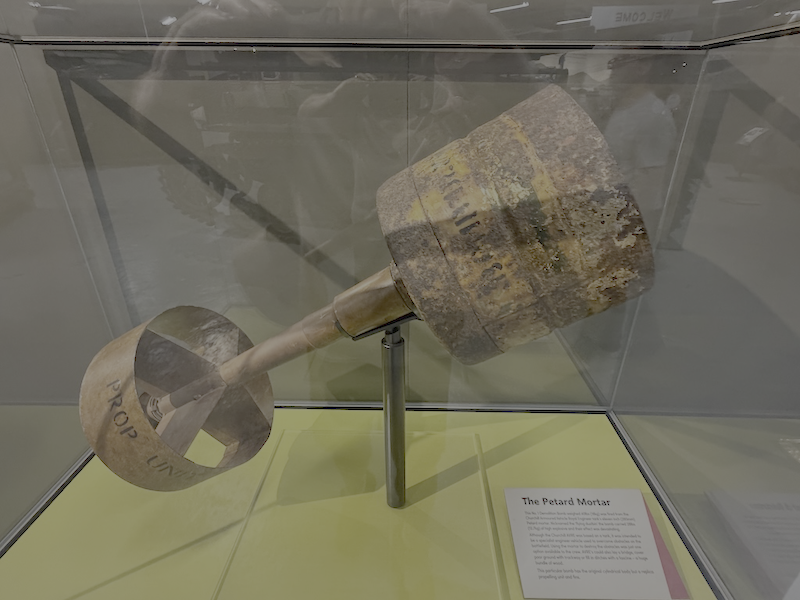 An old, rusty, cylindrical projectile or mortar shell displayed in a glass case at a museum with a label explaining its history.