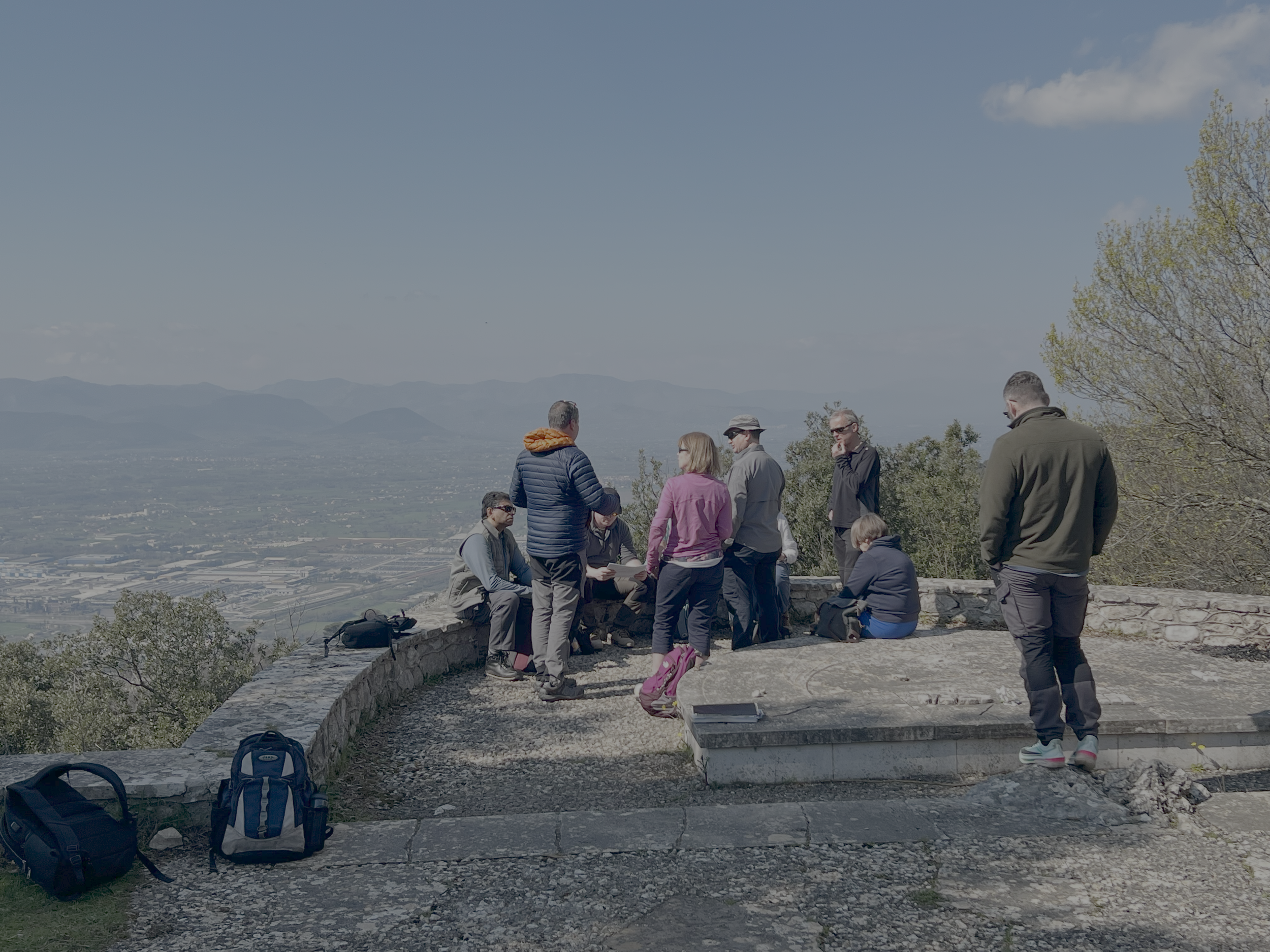 A group of people standing and sitting on a stone ledge at an outdoor viewpoint overlooking a valley with mountains in the distance. Some are engaged in conversation, and backpacks are placed on the ground nearby. A few backpacks are visible in the foreground.