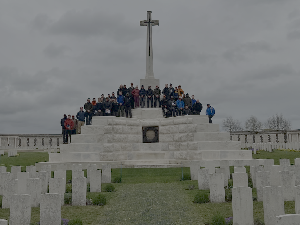 Group of people standing on a white memorial with a cross on top, surrounded by numerous white headstones in an outdoor cemetery under cloudy sky.