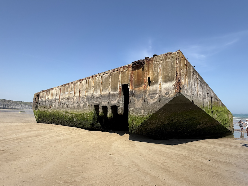 A large, abandoned concrete shipwreck on a sandy beach with some moss and algae growth on its hull, under a clear blue sky.