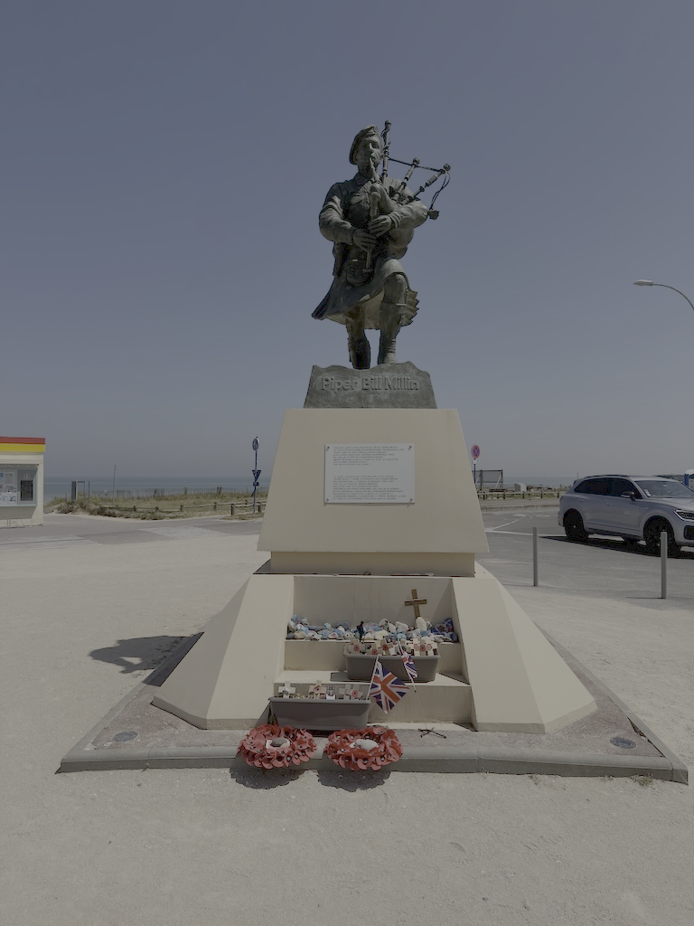 Statue of a soldier playing bagpipes on a pedestal, with a plaque and memorial items including flowers and Union Jack flags in front, set outdoors near the beach.