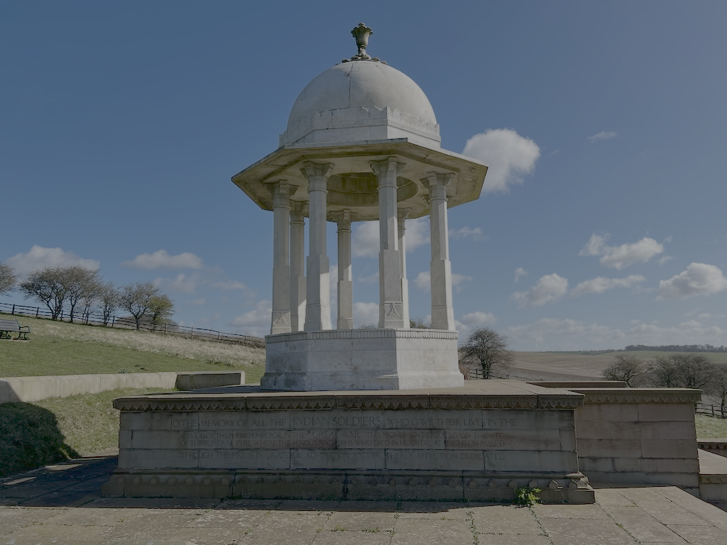 White memorial structure with a domed roof, supported by columns, situated outdoors on a platform, with open sky and sparse trees in the background.