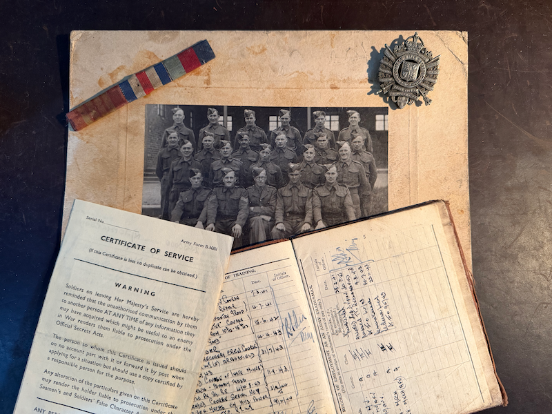 Black and white photograph of soldiers in military uniforms, likely from World War II, displayed on an aged document paper, with a military medal ribbon, a certificate of service, and handwritten notes.