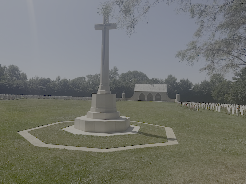 A cemetery with a large stone cross monument in the foreground and rows of smaller white headstones in the background, with trees and a small chapel.