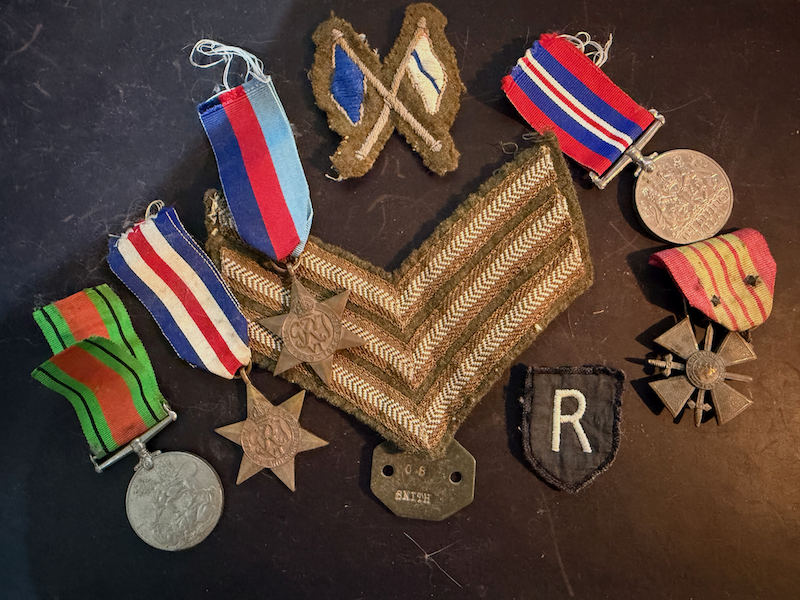 Collection of military medals, patches, and insignia including a military ribbon with a medal, a star-shaped medal, a chevron insignia, and other patches and badges on a dark surface.