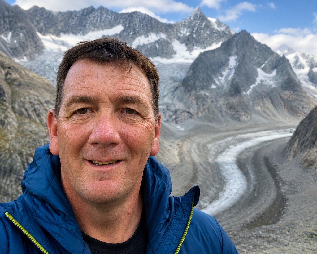 A man in a blue jacket taking a selfie in a mountainous, glacial landscape with snow-capped peaks in the background.