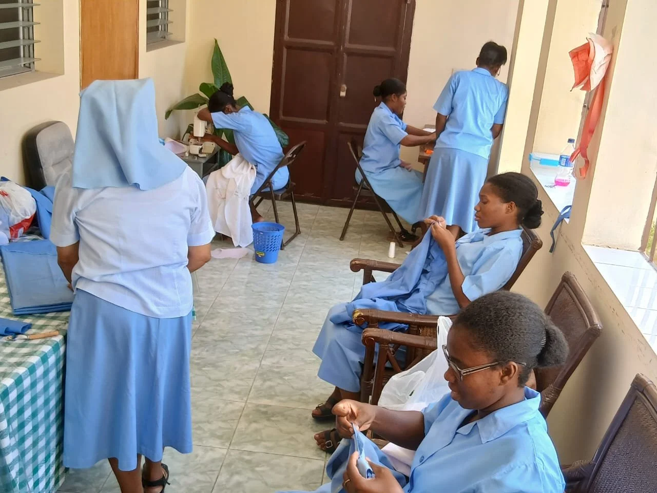 Six Haitian women in light blue uniforms with some wearing head coverings are in a room, engaged in different activities including sewing, folding clothes, and resting. The room has tiled floors, wooden chairs, and a window with metal grills.