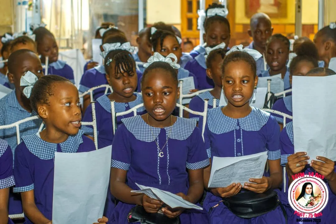 Group of young girls in blue uniforms sitting in a classroom, holding papers, some looking attentive, others appearing confused or engaged.