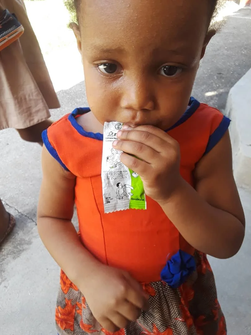 A young child with short hair wearing an orange sleeveless shirt with blue trim, eating a bag of Nouri Mamba / Plumpy sup. 