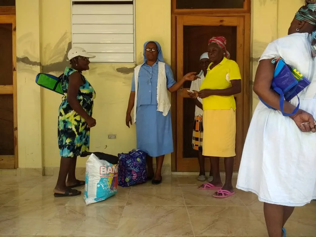 Sr Judith is overseeing a community Food distribution at the Little Sisters Baradères Hospital with 3 women standing and talking inside a building with a yellow wall and wooden door. One woman is wearing a floral dress and hat, carrying a shelter bag