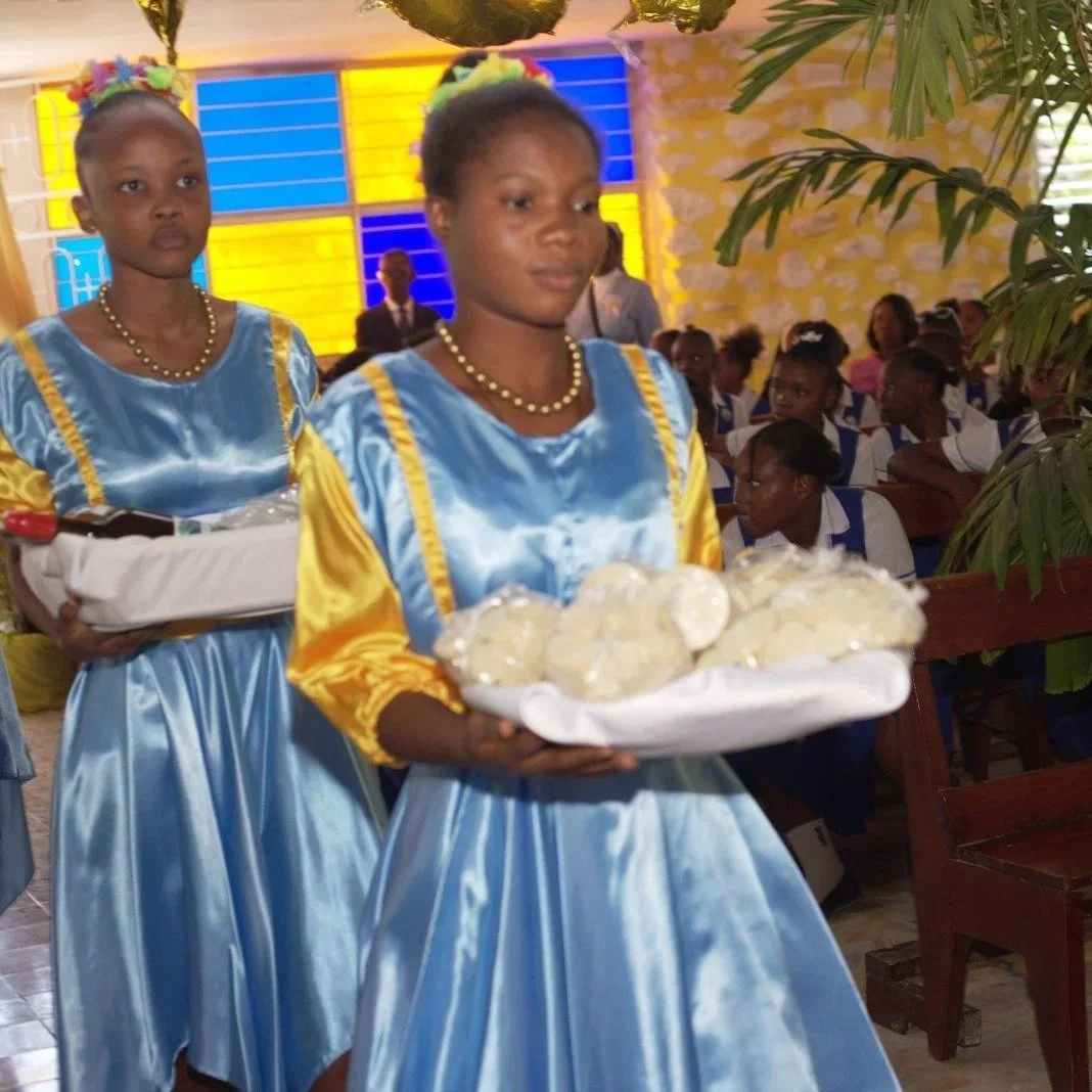 Two young girls in blue and yellow dresses with pearl necklaces, holding trays with white round baked goods in a vibrant indoor church setting, with an audience seated in the background.