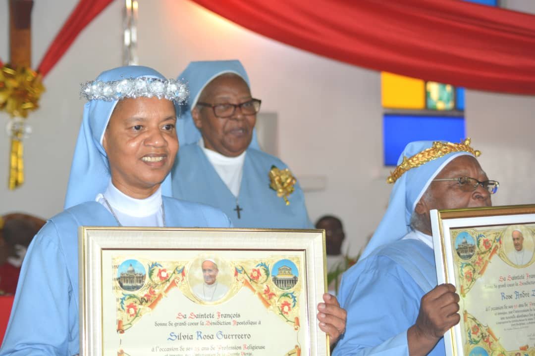 Three nuns dressed in blue holding framed certificates during a religious event with red and gold decorations.