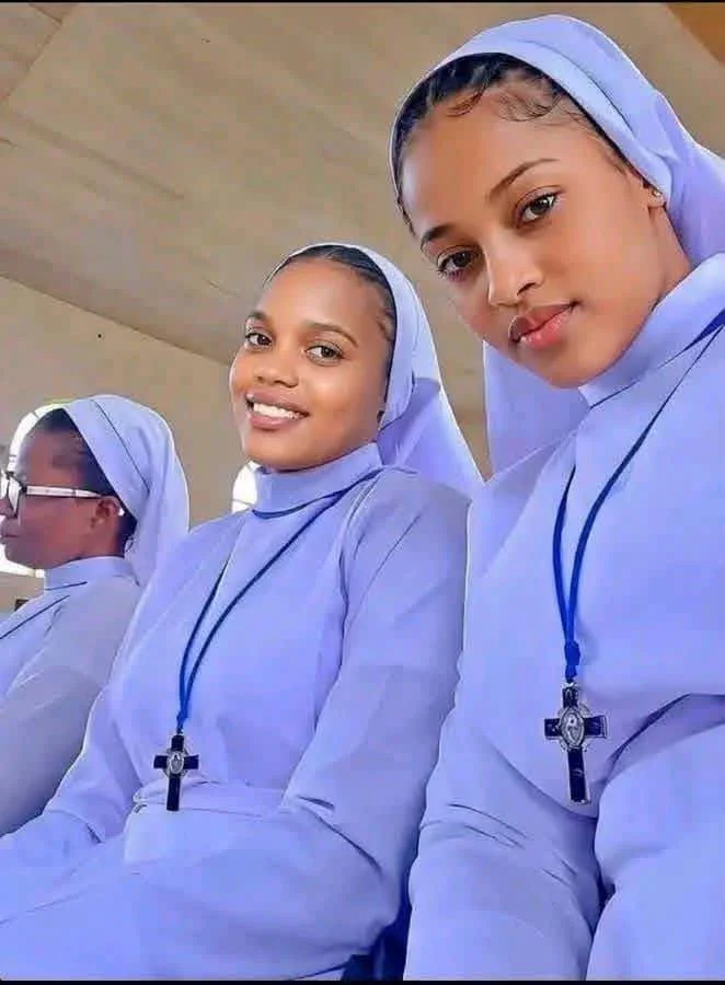 Three nuns dressed in white habits with blue head coverings, sitting in a church or chapel. The two women in the foreground are smiling and wearing crosses around their necks.