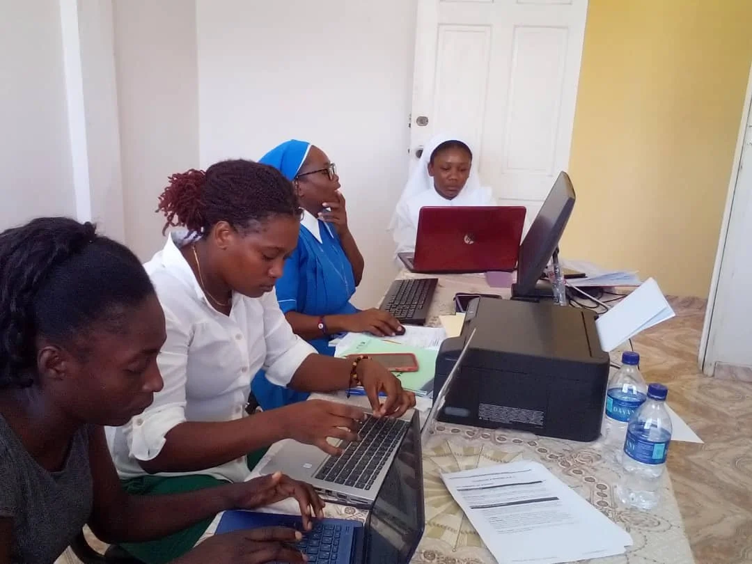 Four women seated at a table working on laptops, with papers and water bottles in a room with a yellow wall and a closed door.