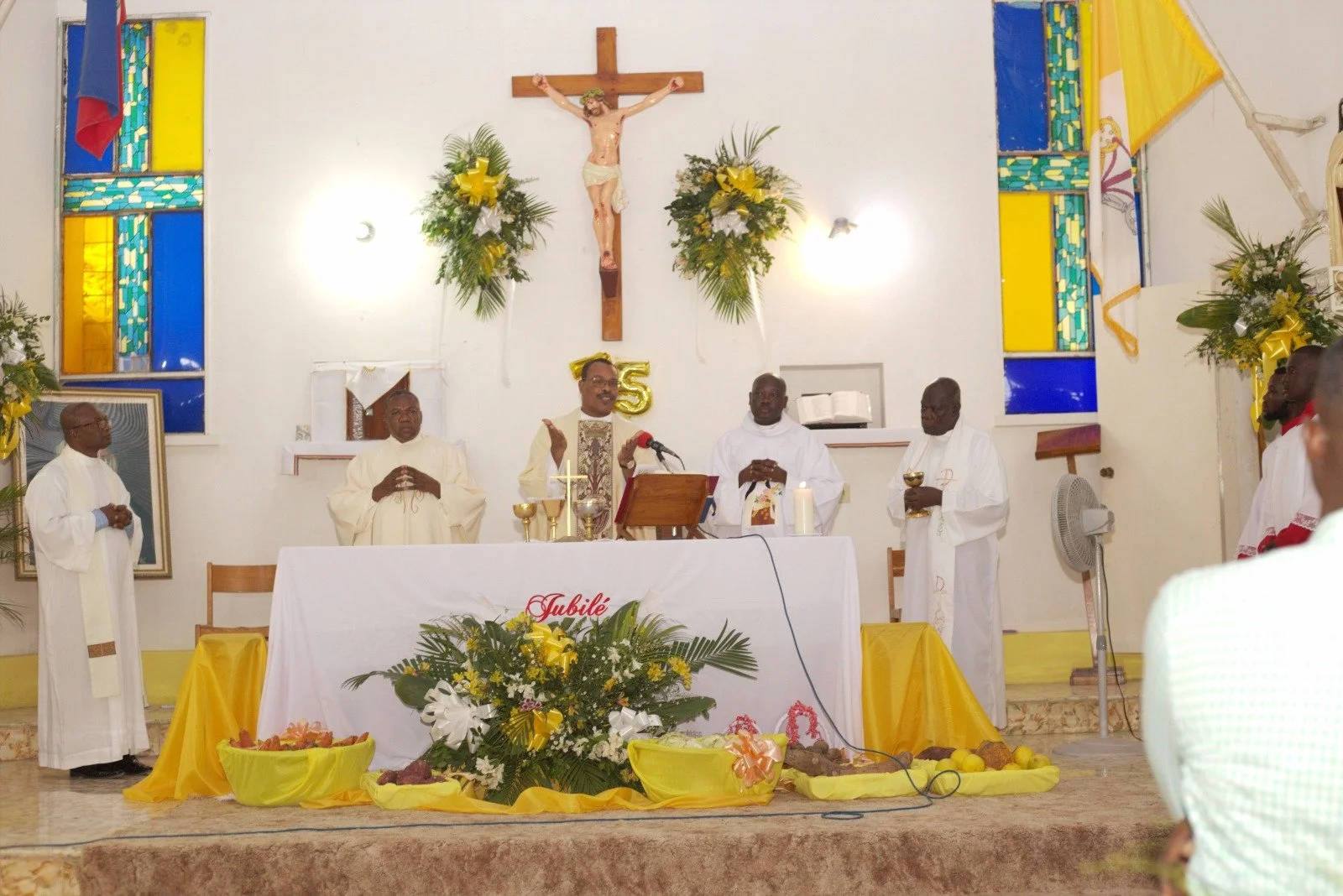 A church service with priests and clergy celebrating at the altar decorated with yellow and white flowers, with a crucifix and colorful stained glass windows in the background.