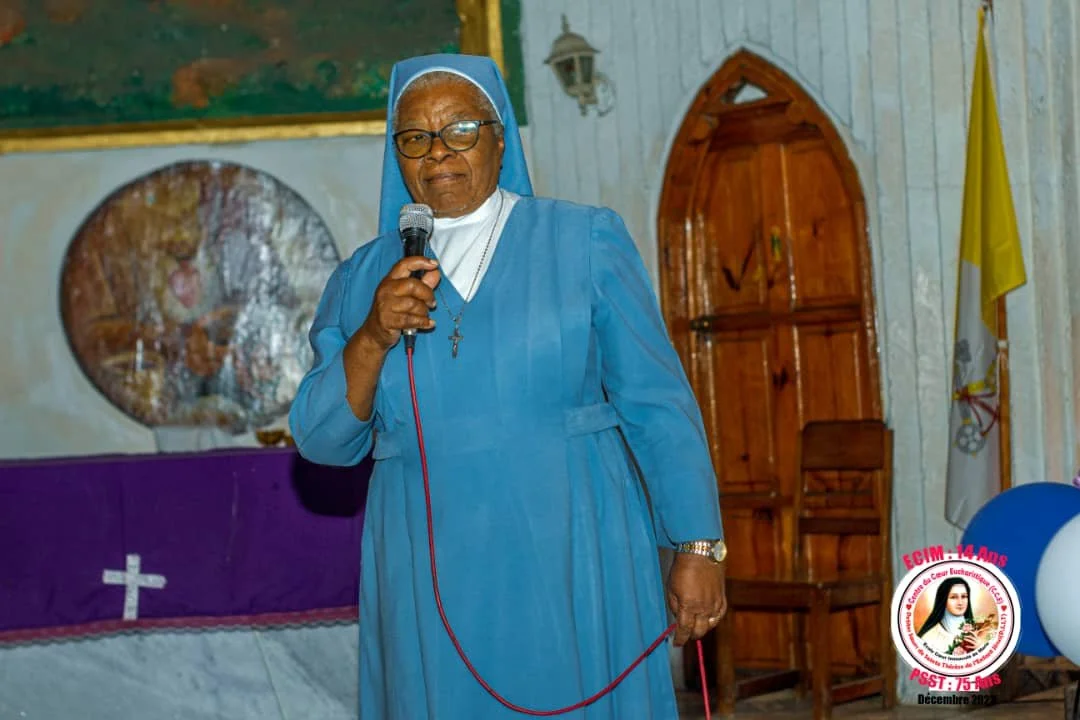 A nun in blue habit and glasses holding a microphone in a church setting.